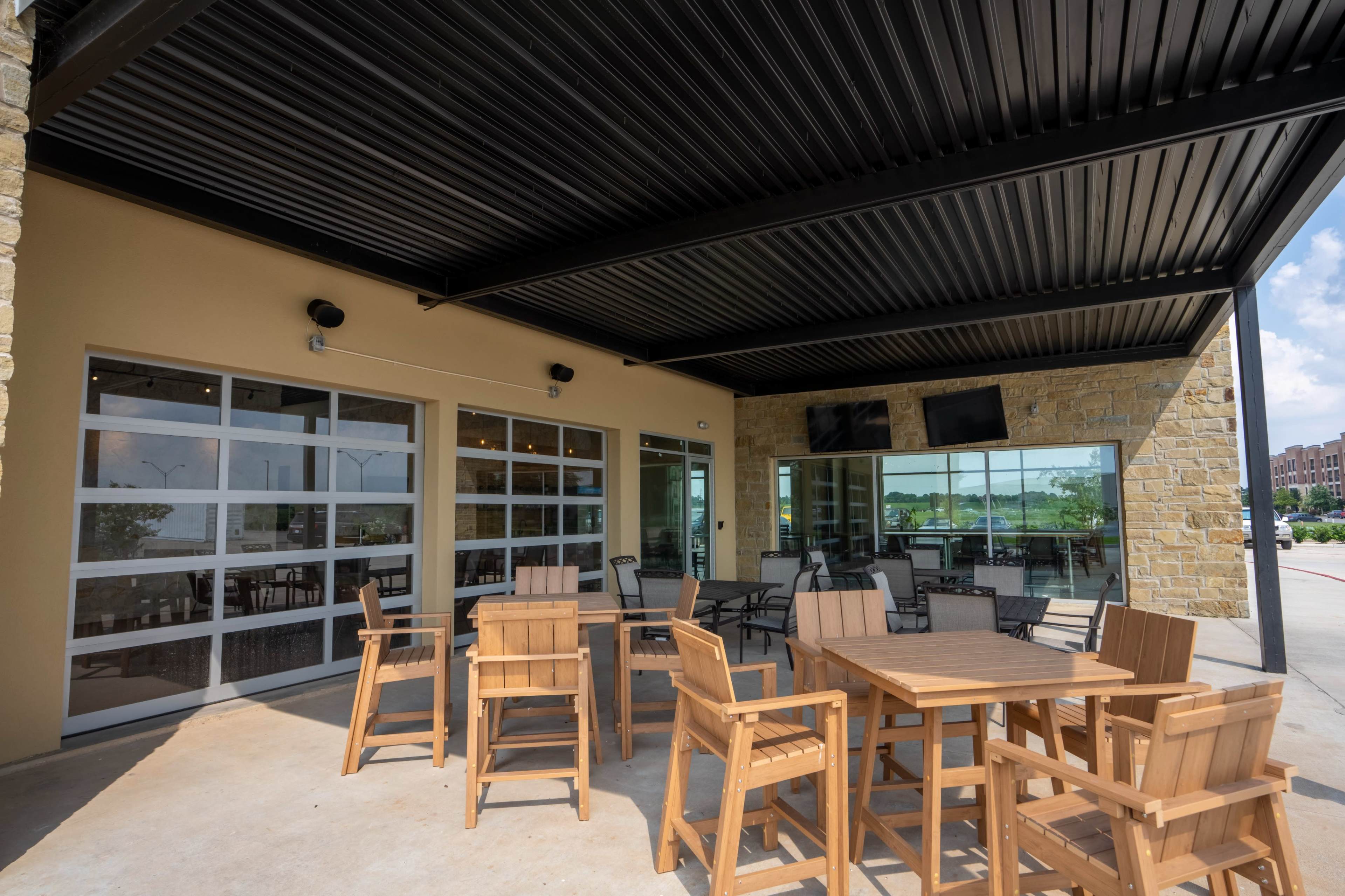An outdoor seating area with wooden furniture under a covered patio, adjacent to a building with large glass doors.