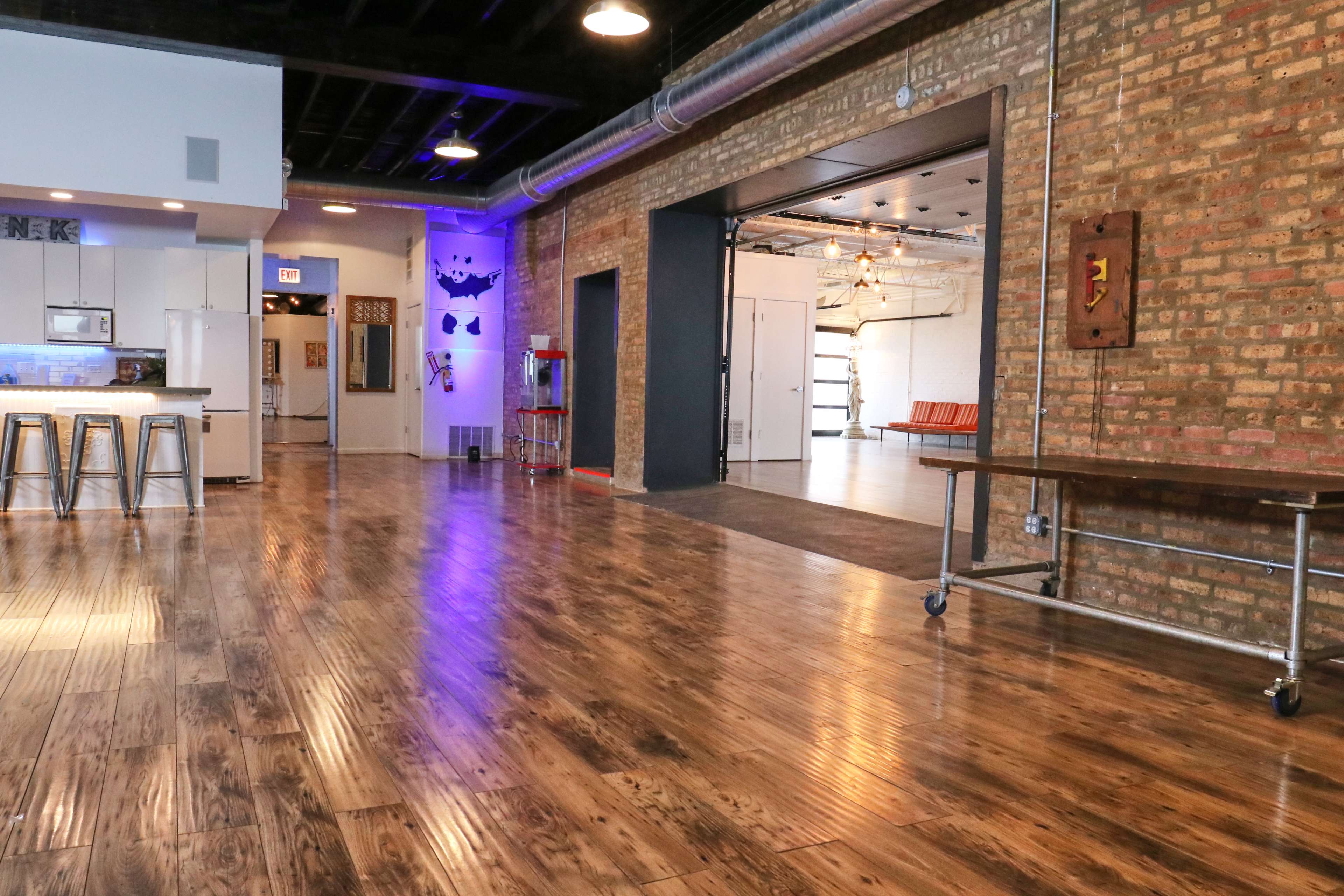The image shows a spacious industrial-style room with wooden floors, exposed brick walls, and a kitchenette on one side.