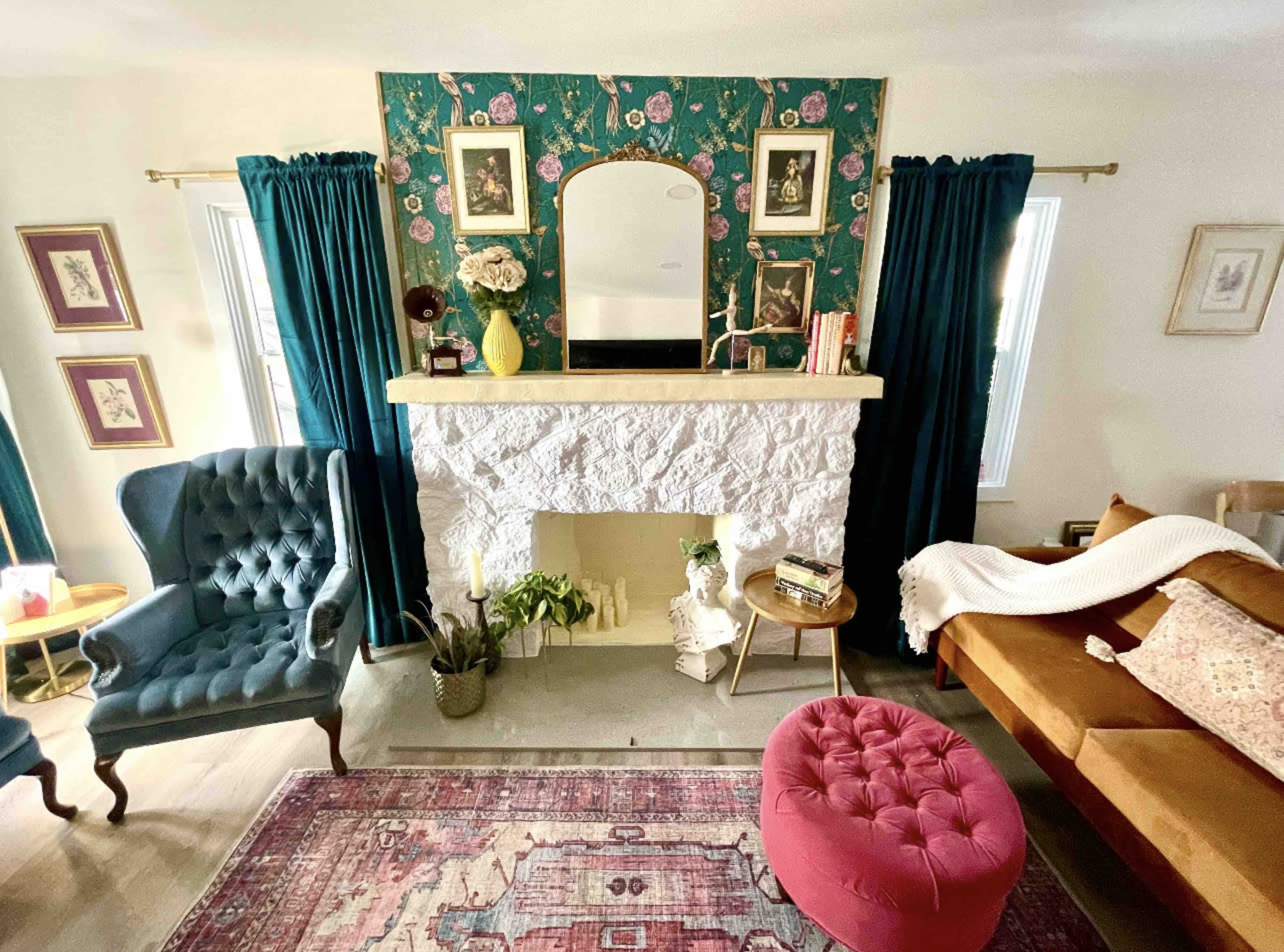 A living room with a stone fireplace, faux floral wallpaper, and a mix of vintage furniture, including a blue armchair and a brown sofa.