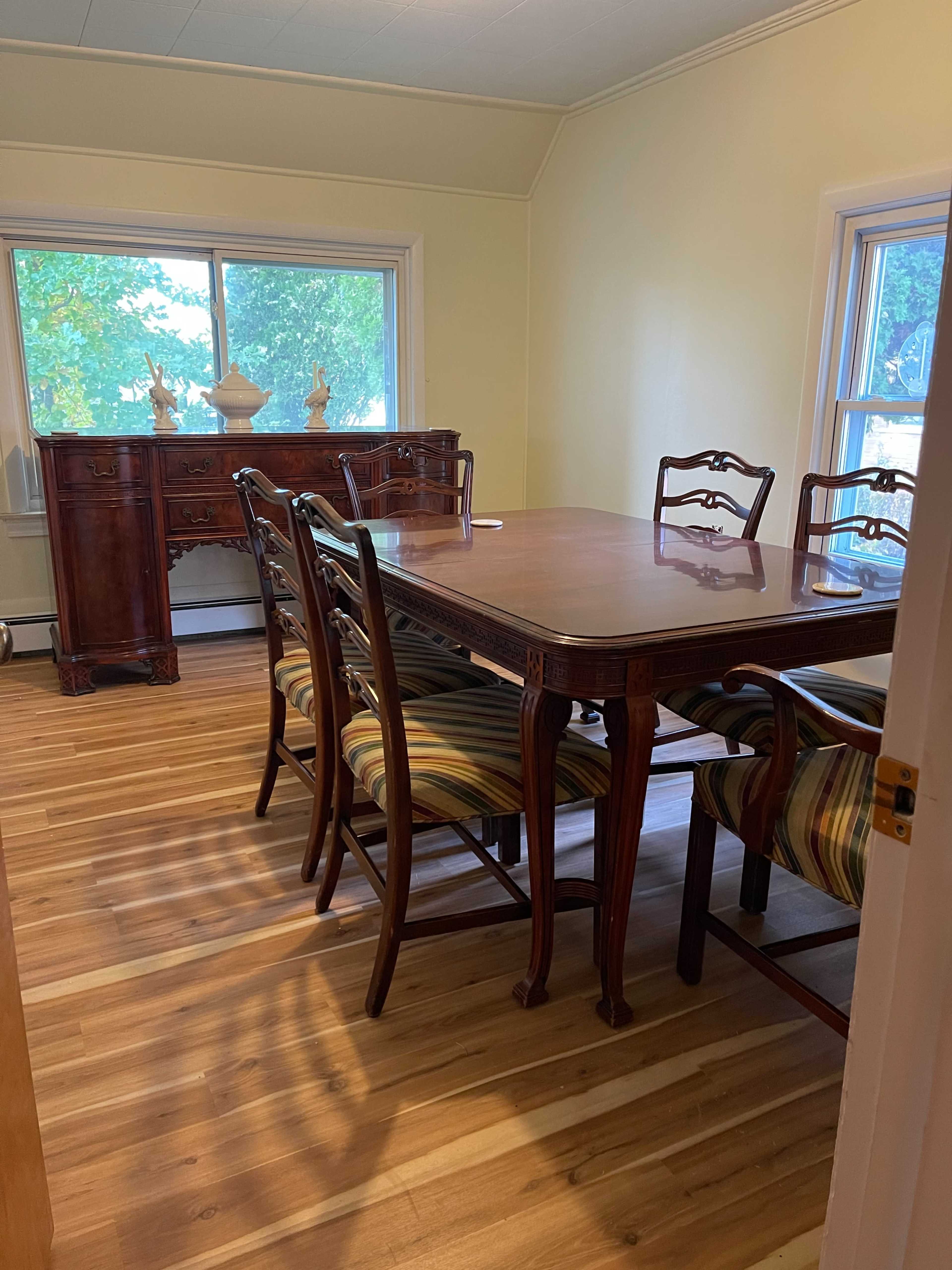 A dining room features a wooden table surrounded by six chairs, with a sideboard against the wall and large windows letting in natural light.