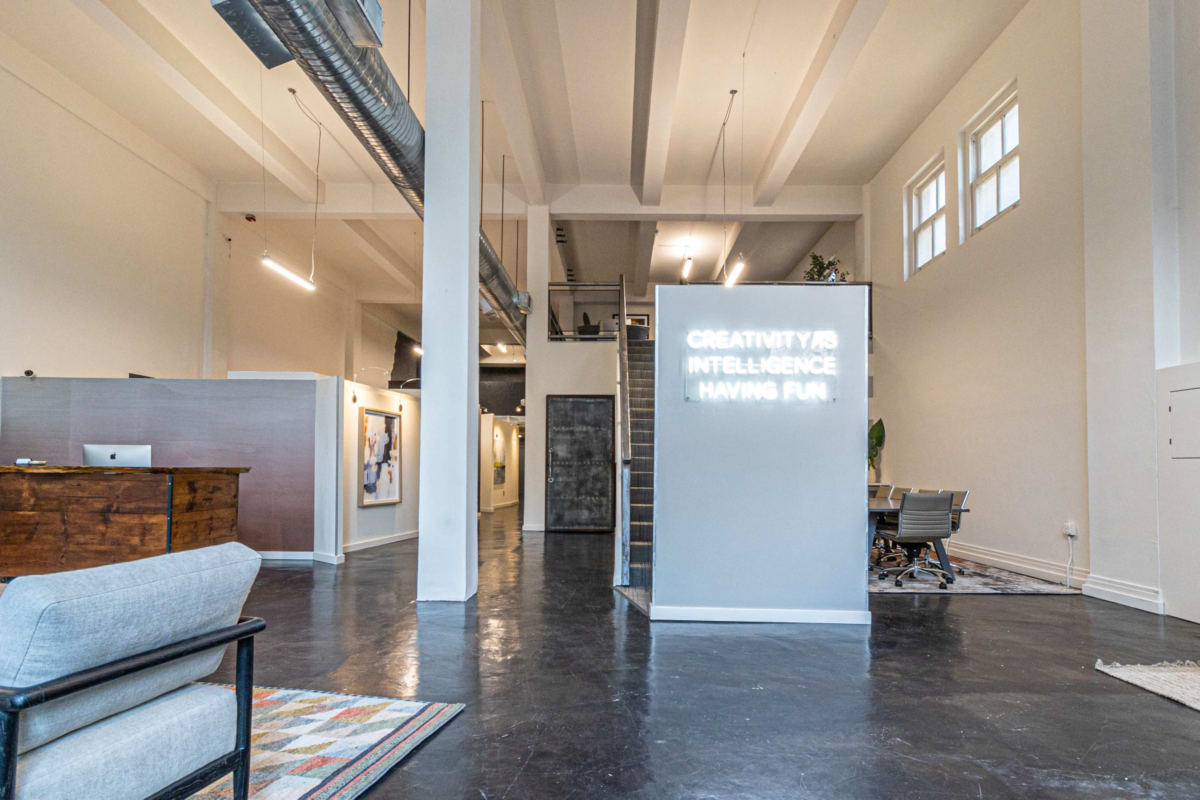 The image shows a spacious, modern office interior featuring a reception area with a wooden desk, a meeting space with a round table, and a wall displaying a glowing sign.