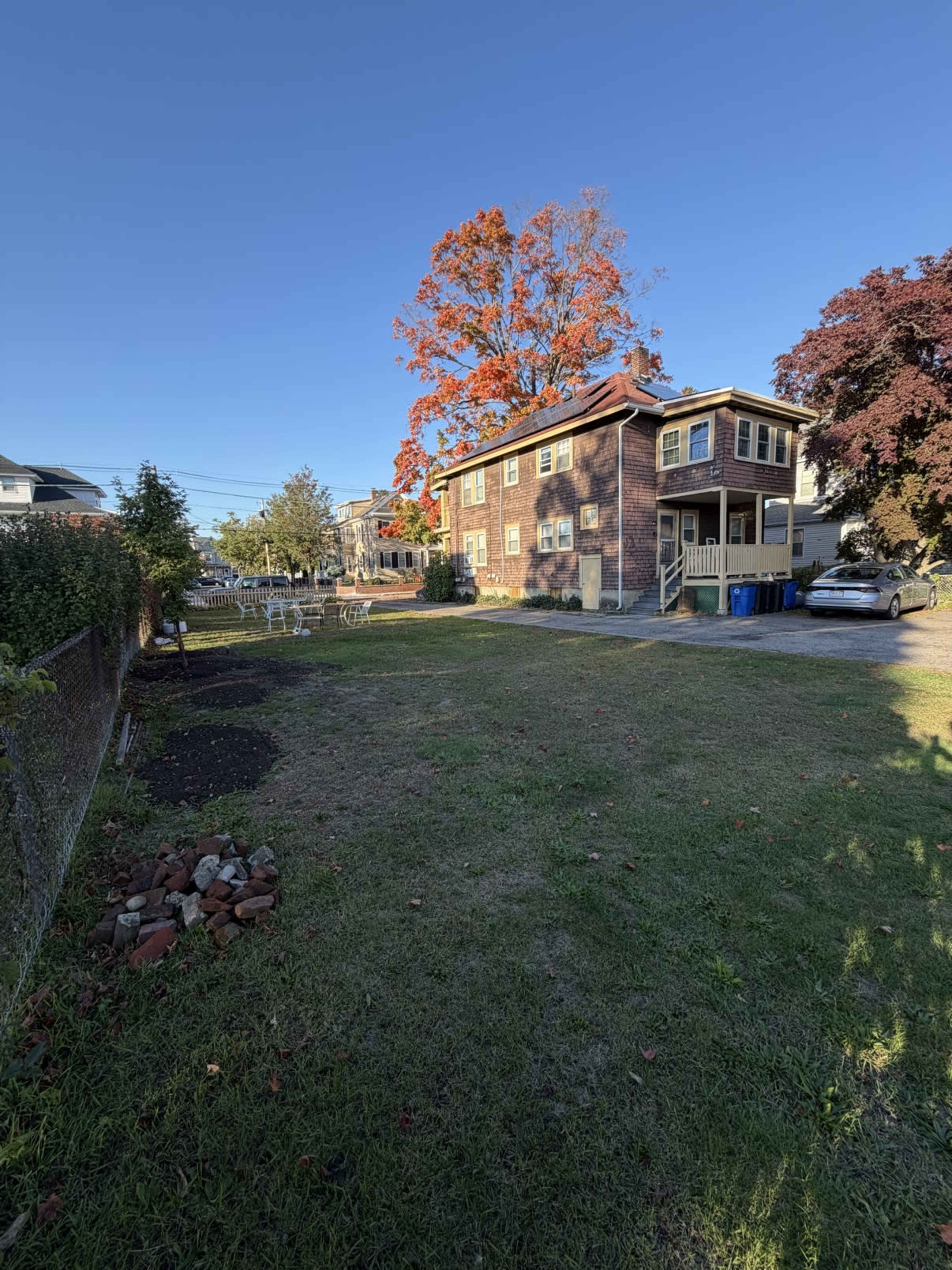 The image shows a two-story brick house surrounded by a grassy yard, with colorful trees in the background and some outdoor furniture in the distance.