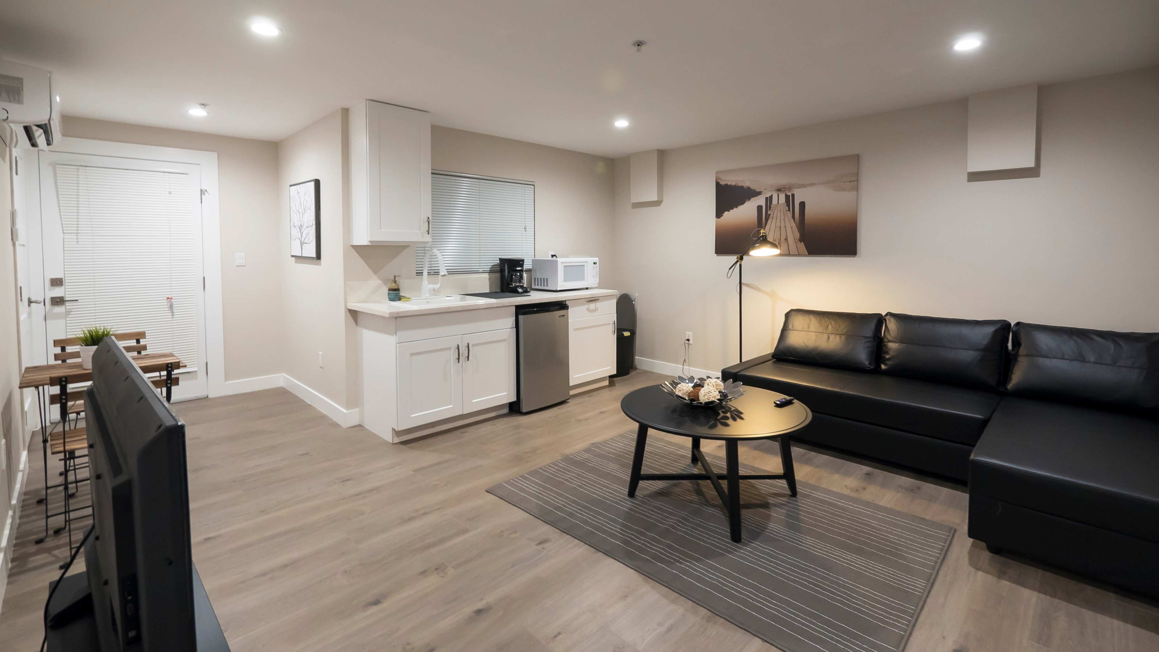 A modern living area with a black sectional sofa, a round coffee table, a television, and a kitchenette featuring a microwave and a refrigerator.