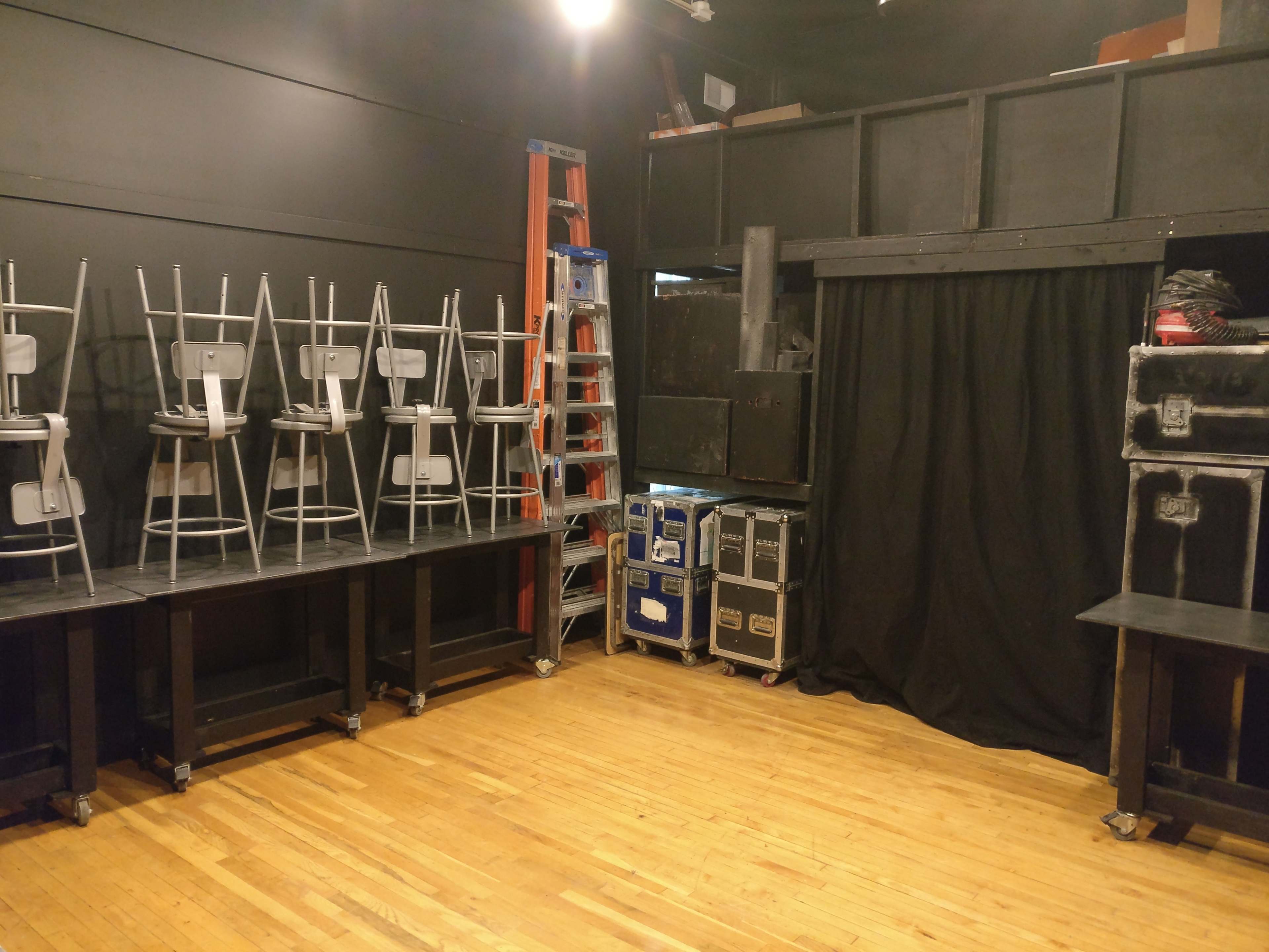 A dimly lit room with stacked chairs on tables, a ladder, and various equipment cases against the walls.