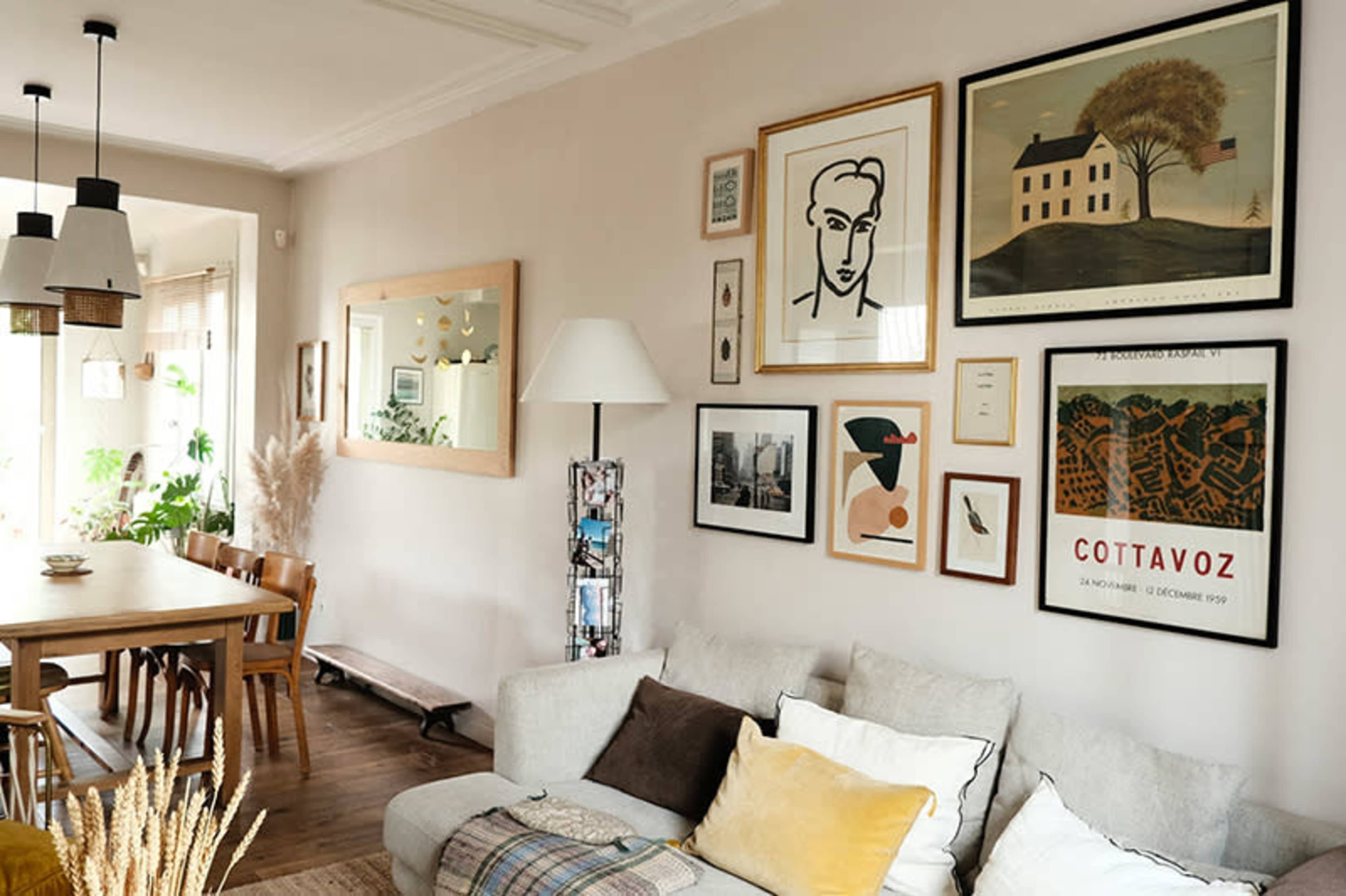 The image shows a cozy living room with a sectional sofa adorned with pillows, a dining table set nearby, and a wall featuring various framed artworks.