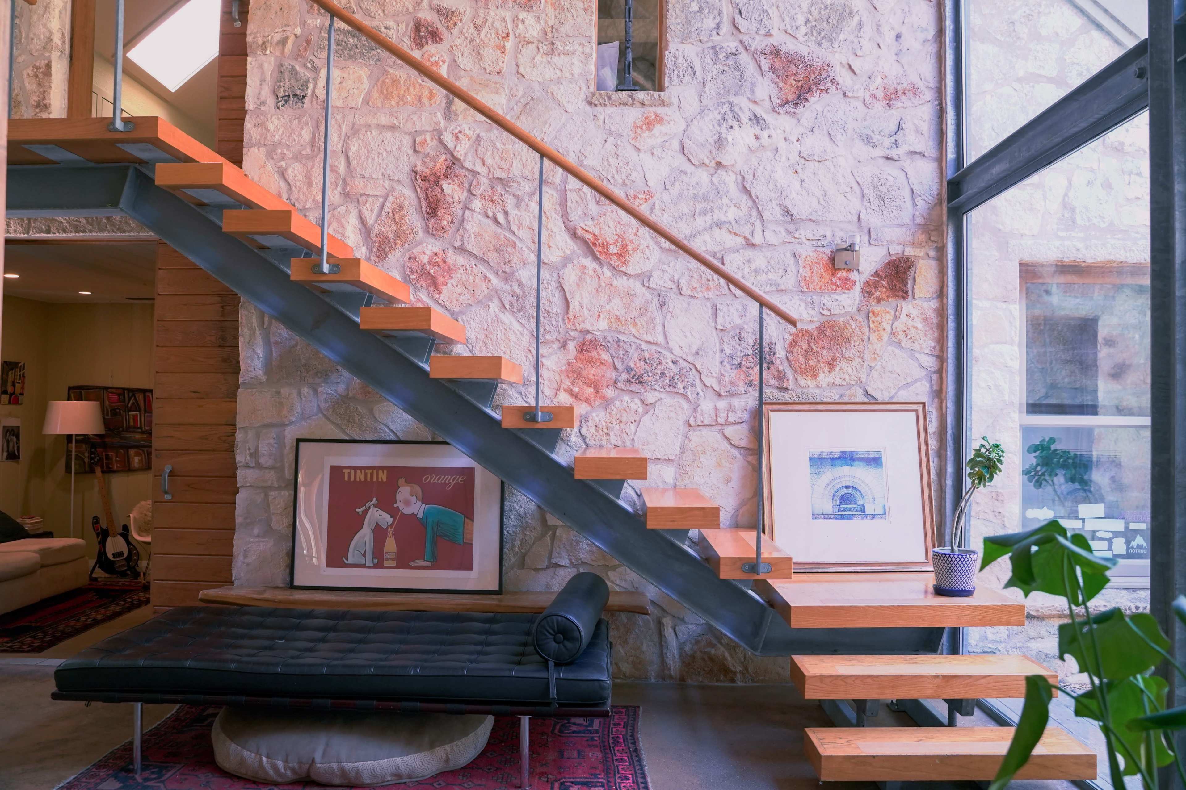 A wooden staircase with metal railings ascends beside a stone wall, featuring framed artwork and a plant on a side shelf.