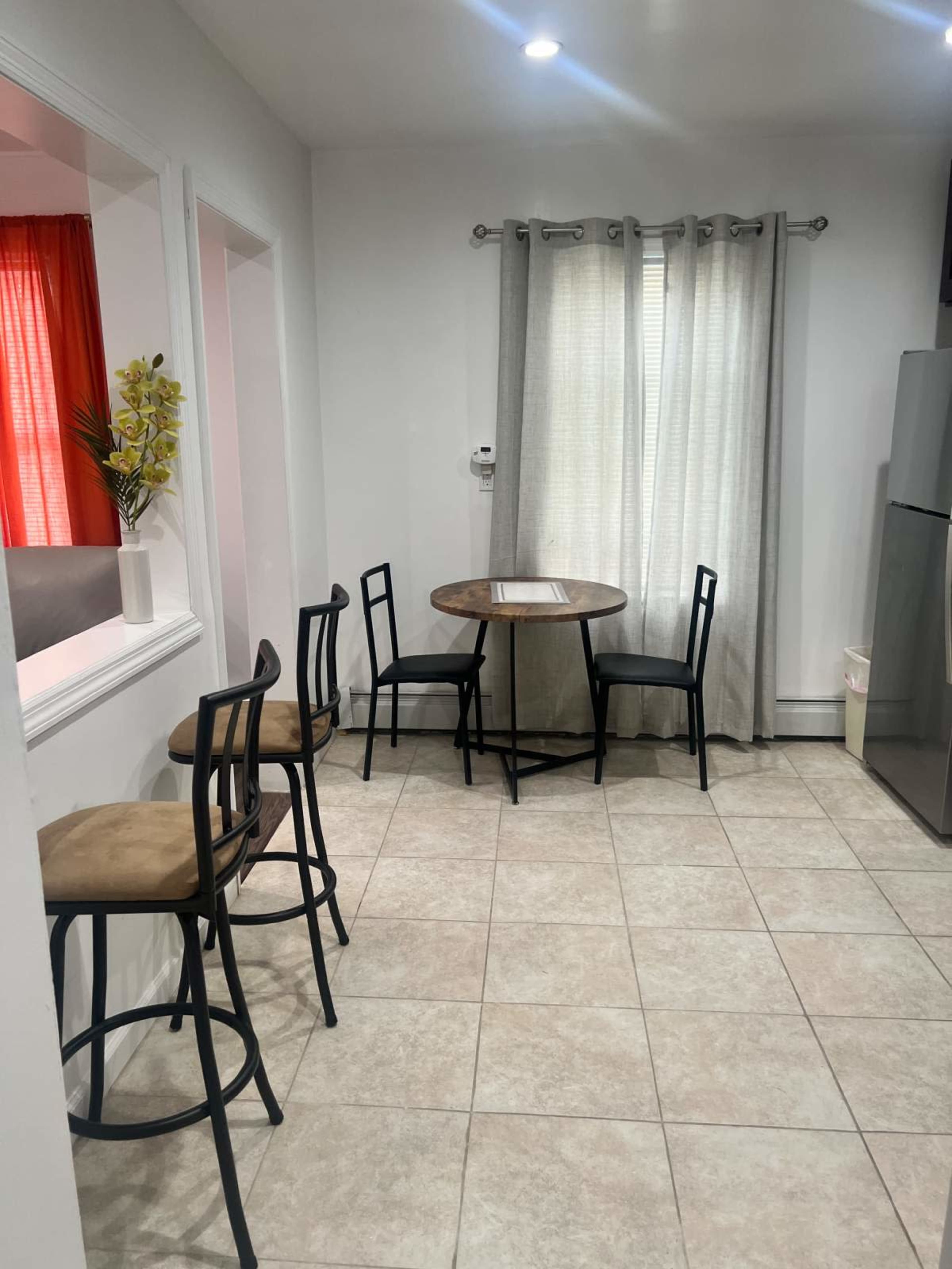 A small dining area features a round table surrounded by four black chairs and a gray refrigerator in a tiled room with light-colored walls and curtains.