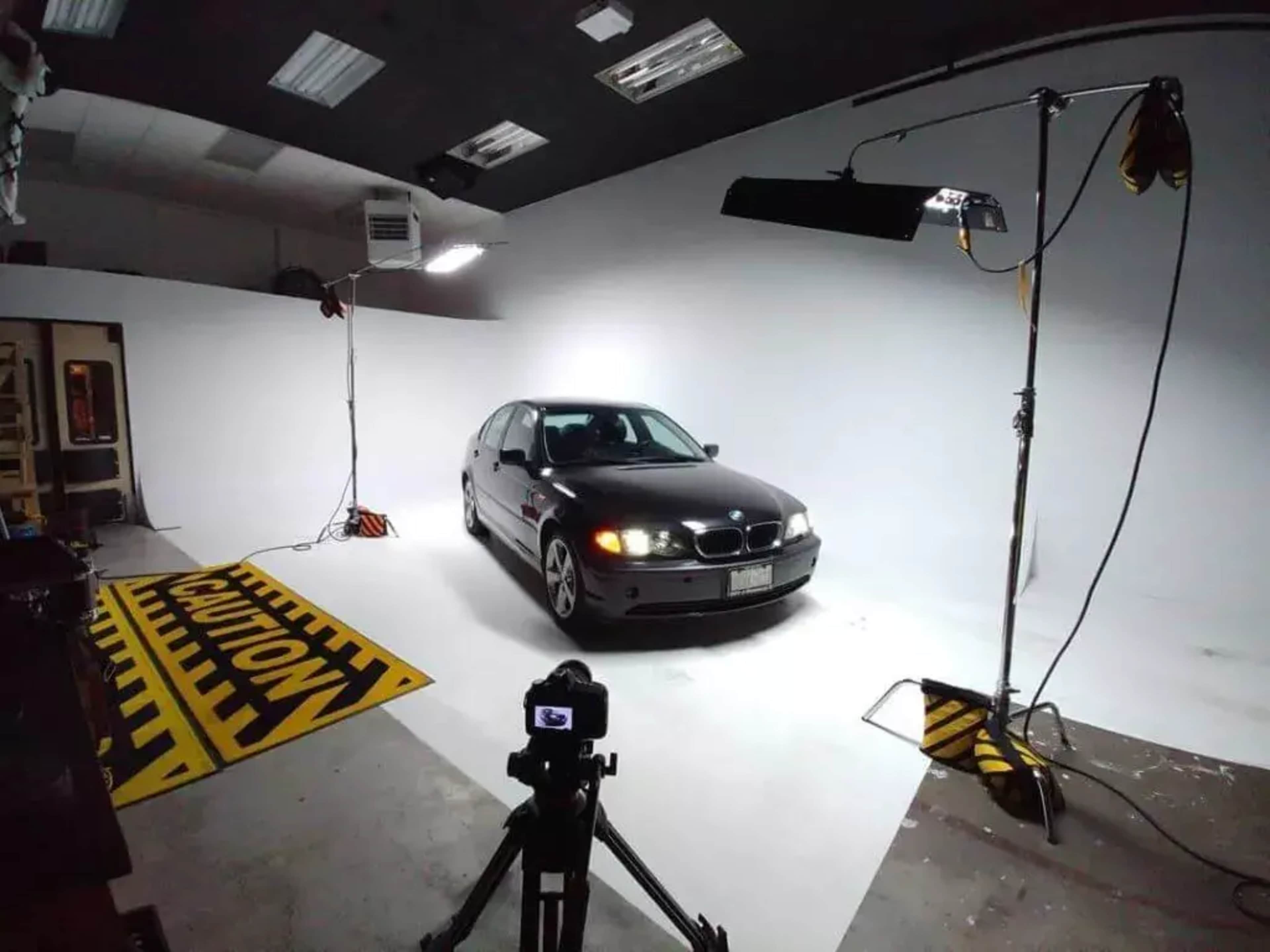 A black BMW sedan is positioned in a well-lit studio against a white backdrop, with lighting equipment and a camera set up for a photoshoot.