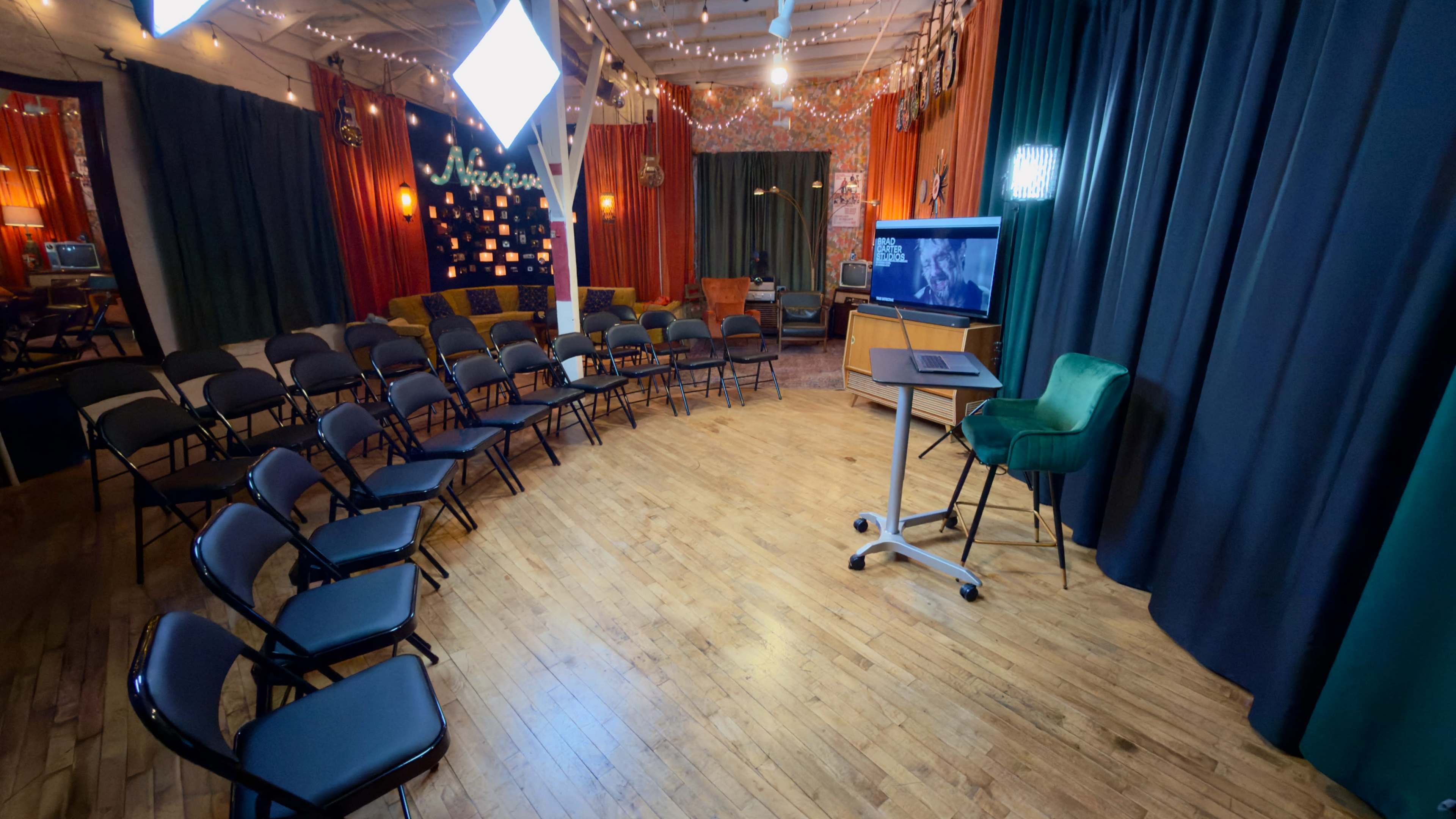 The image shows a well-decorated room set up for a presentation, featuring rows of black chairs, a computer on a mobile stand, and a backdrop adorned with colorful curtains and lights.