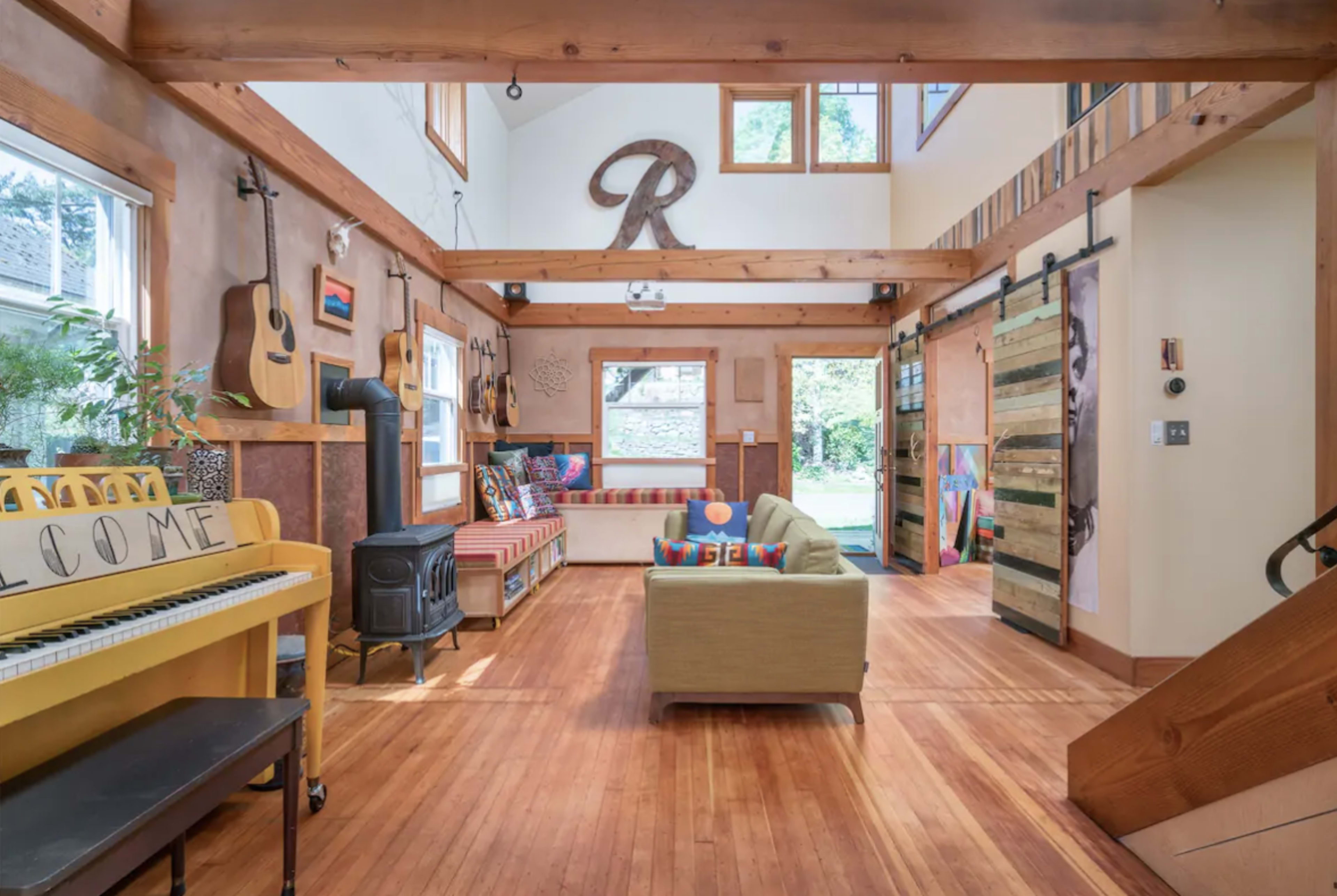 The image shows a brightly lit open living space featuring wooden beams, a piano, seating areas with colorful cushions, and various decorative guitars on the walls.