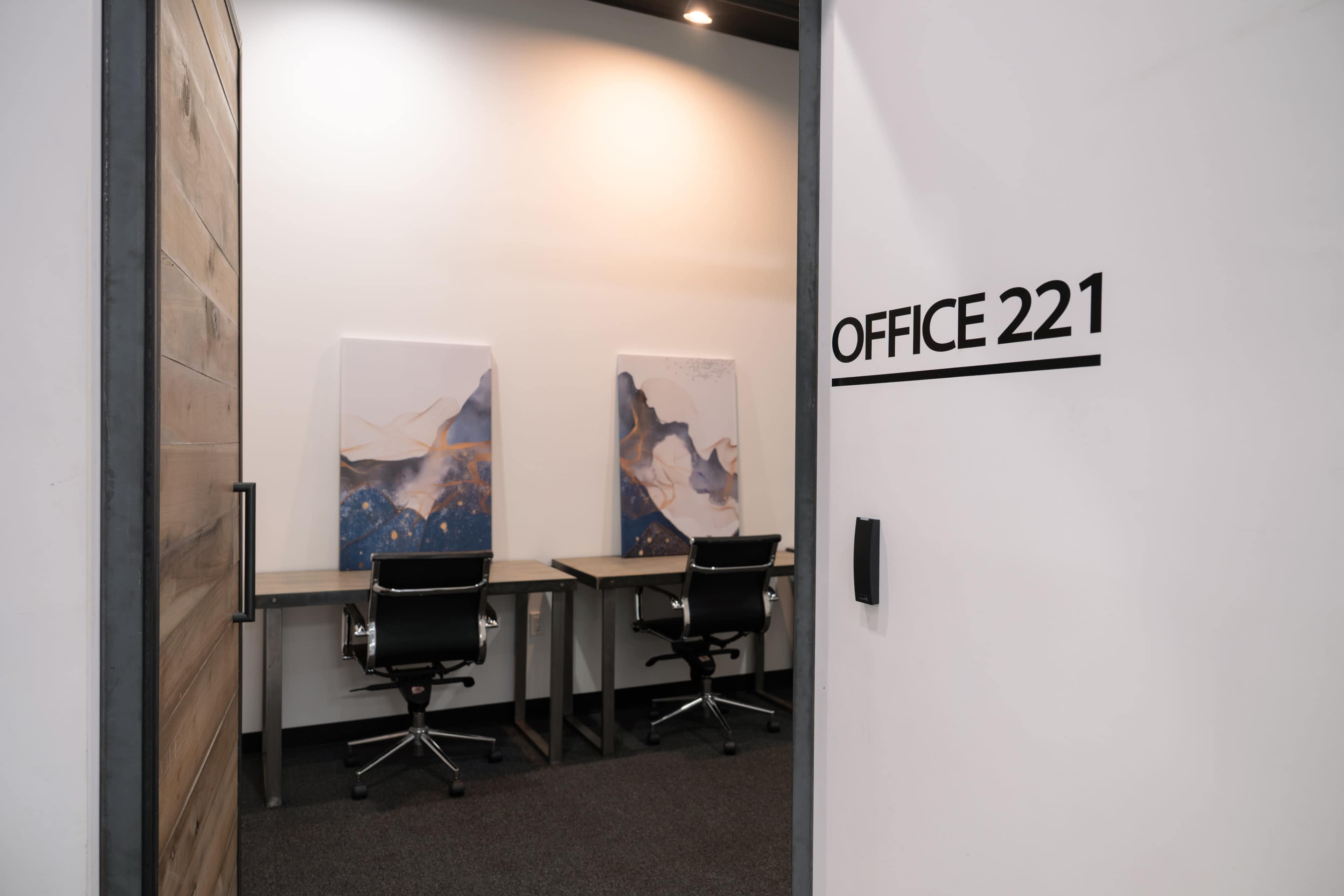 The image shows the entrance to Office 221, featuring two desks with black chairs and abstract artwork on the walls.