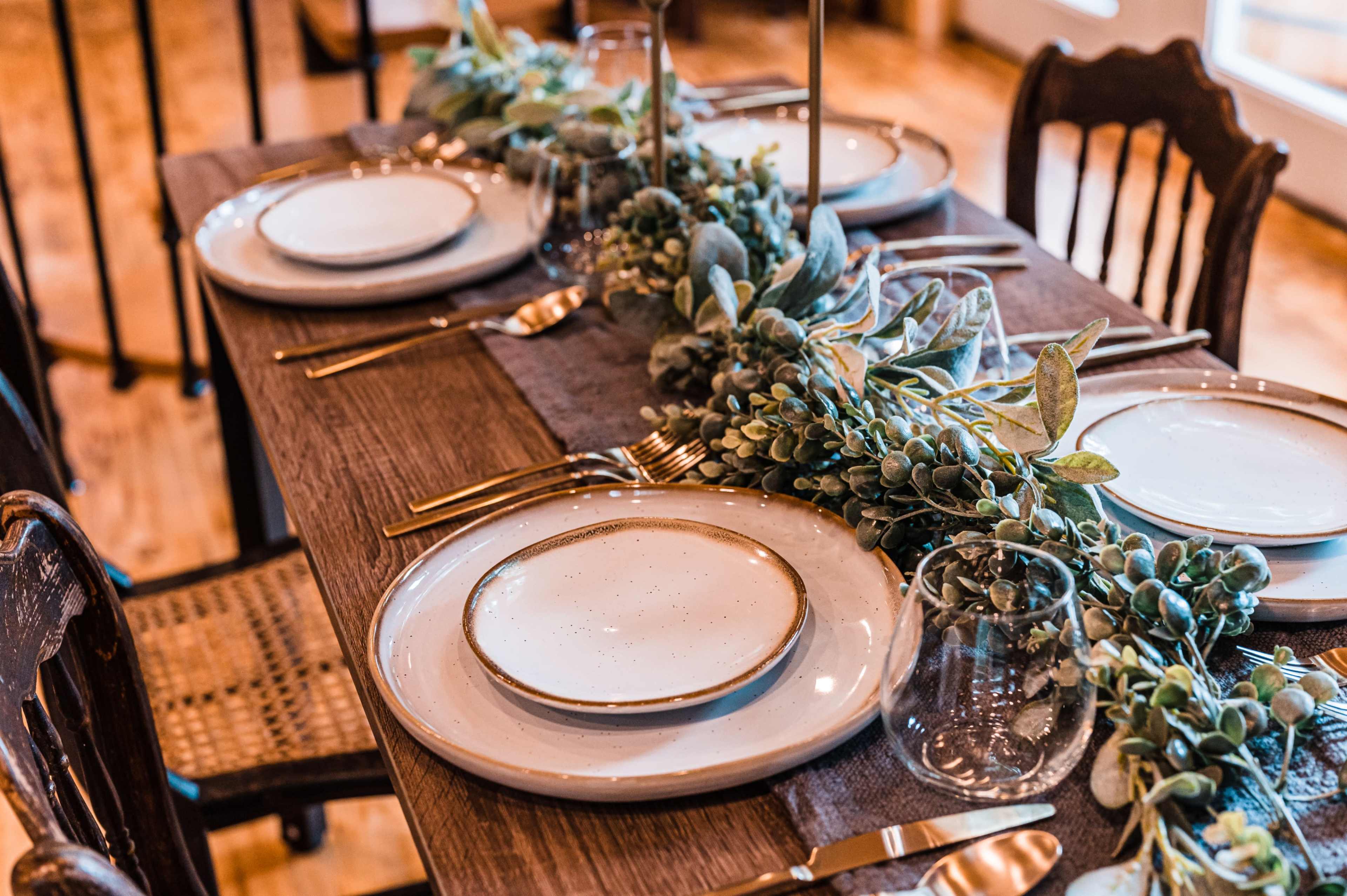 A wooden dining table is set with plates, glassware, and a greenery centerpiece, accompanied by gold utensils.