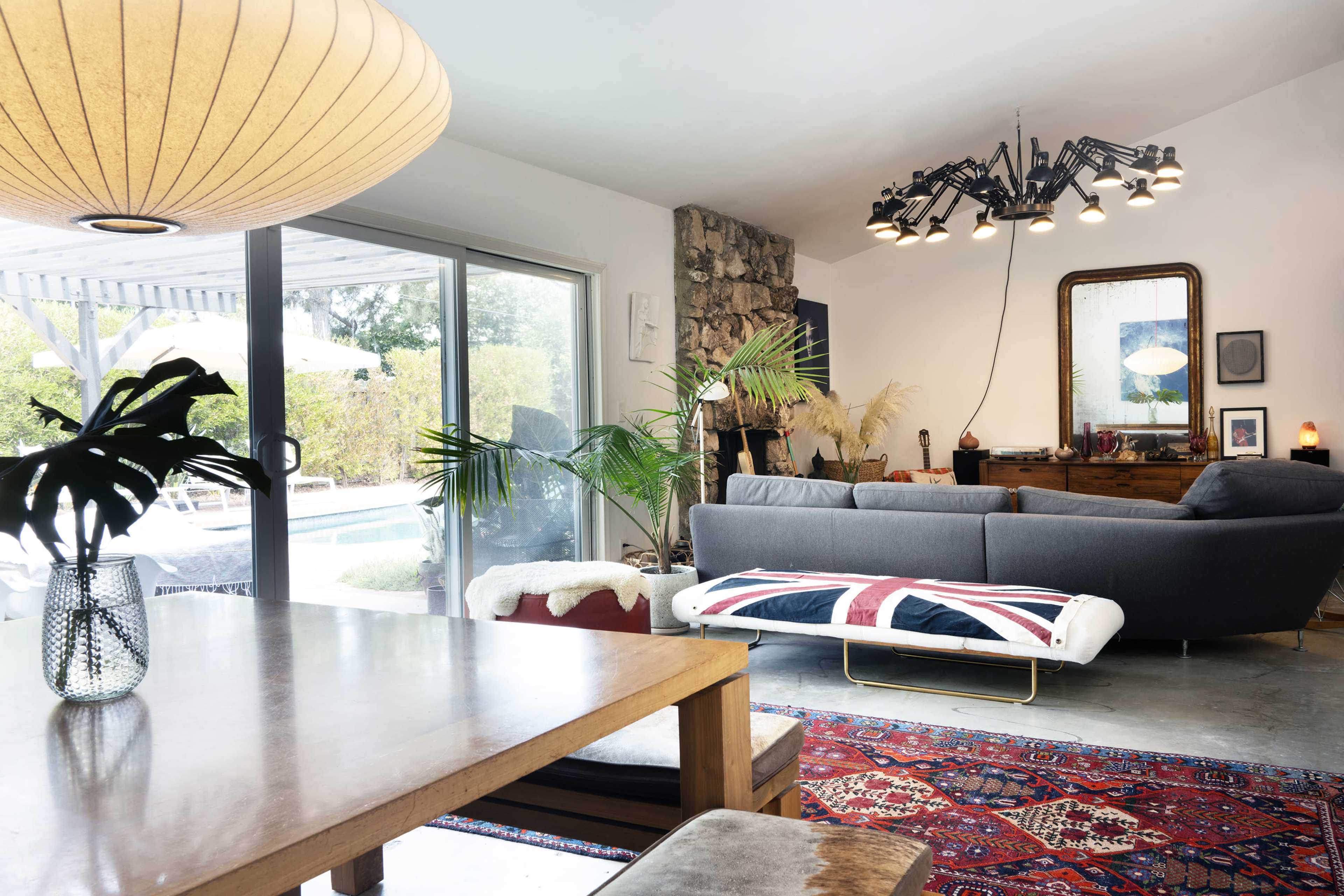 The image shows a modern living room with a gray sofa, a wooden dining table, and large glass doors leading to an outdoor pool area, decorated with plants and a colorful area rug.