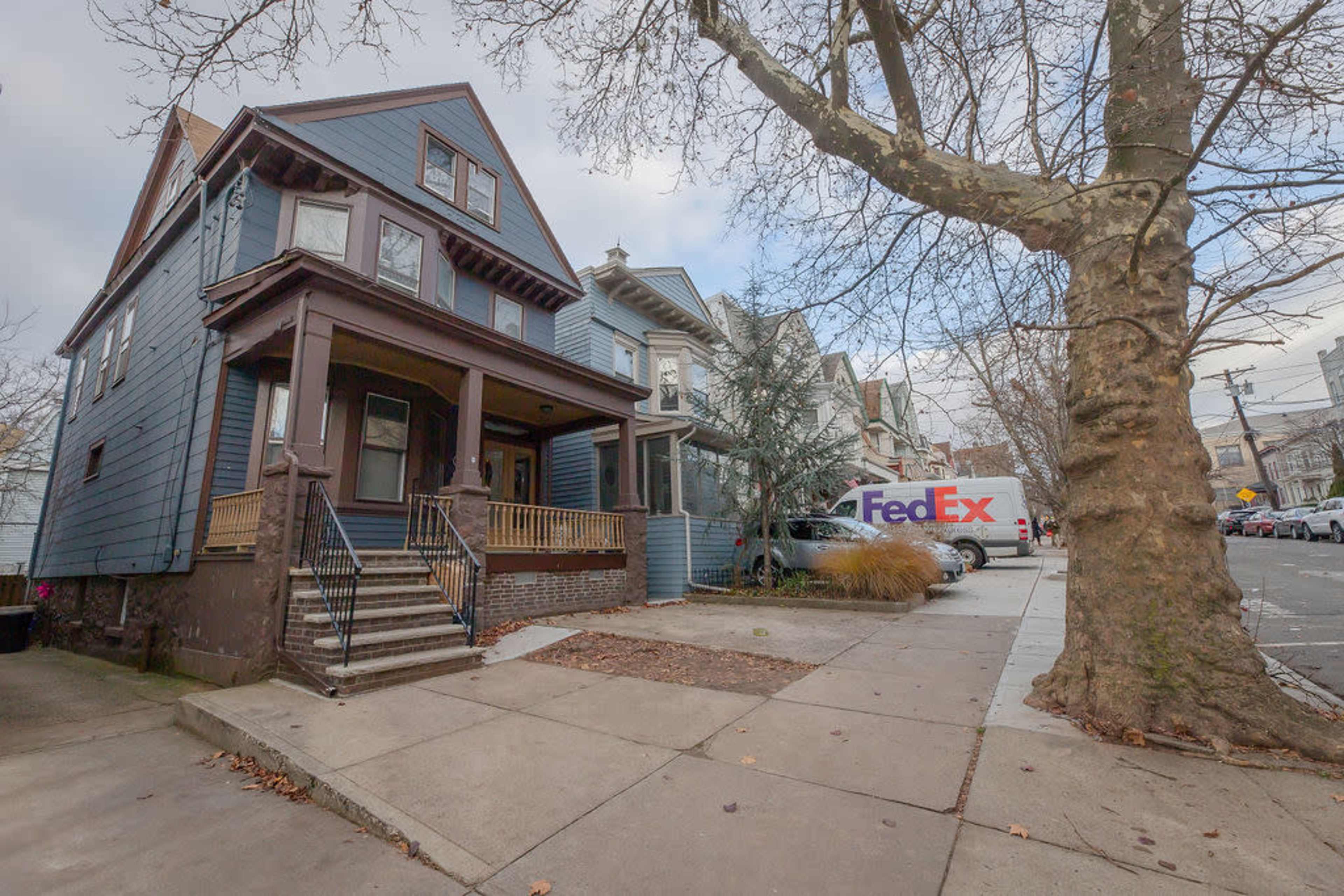 A blue two-story house with a porch stands beside a FedEx delivery truck parked on the street in an urban neighborhood.