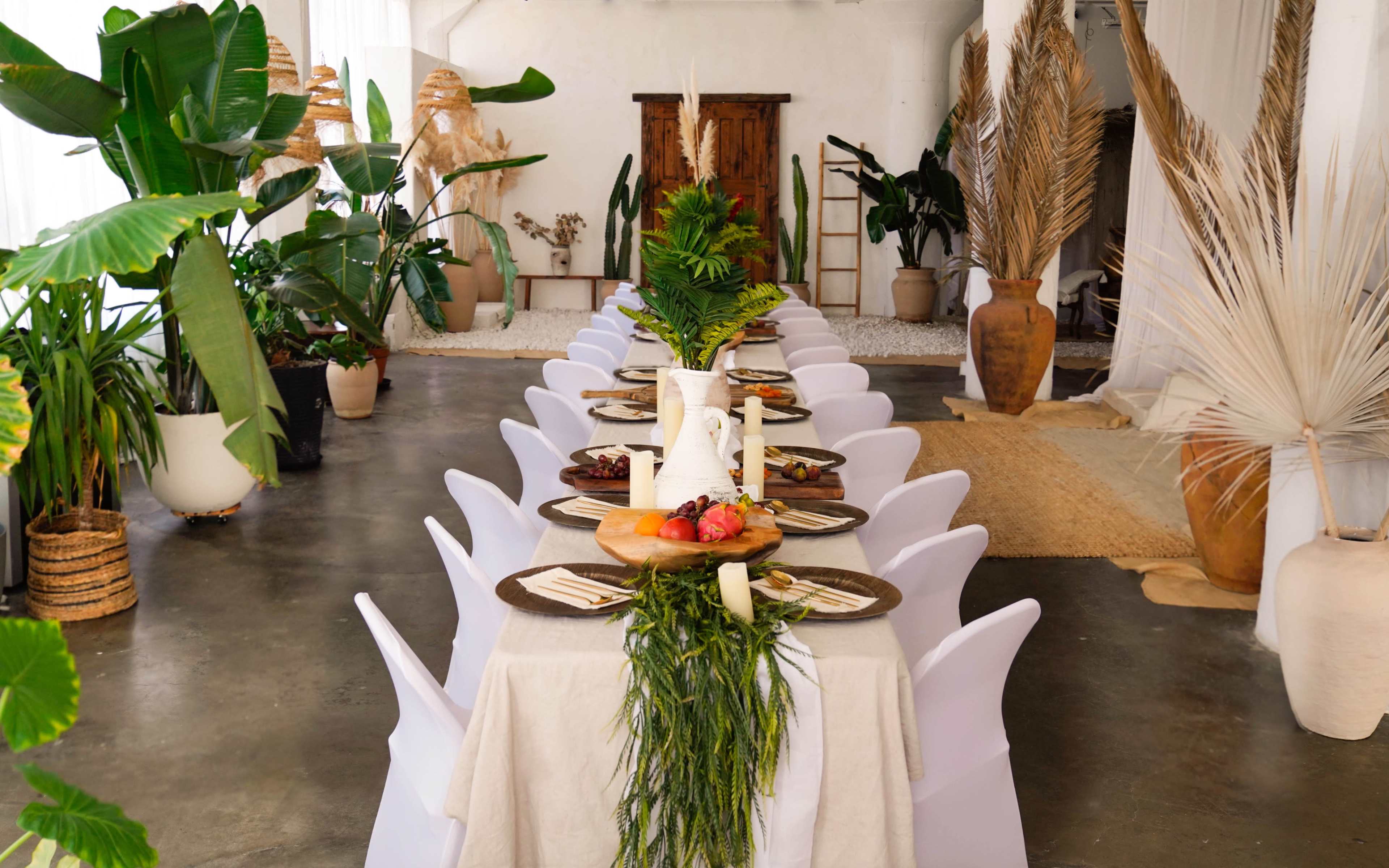 A long dining table is set with plates and decorative elements, surrounded by potted plants in a spacious, modern indoor environment.