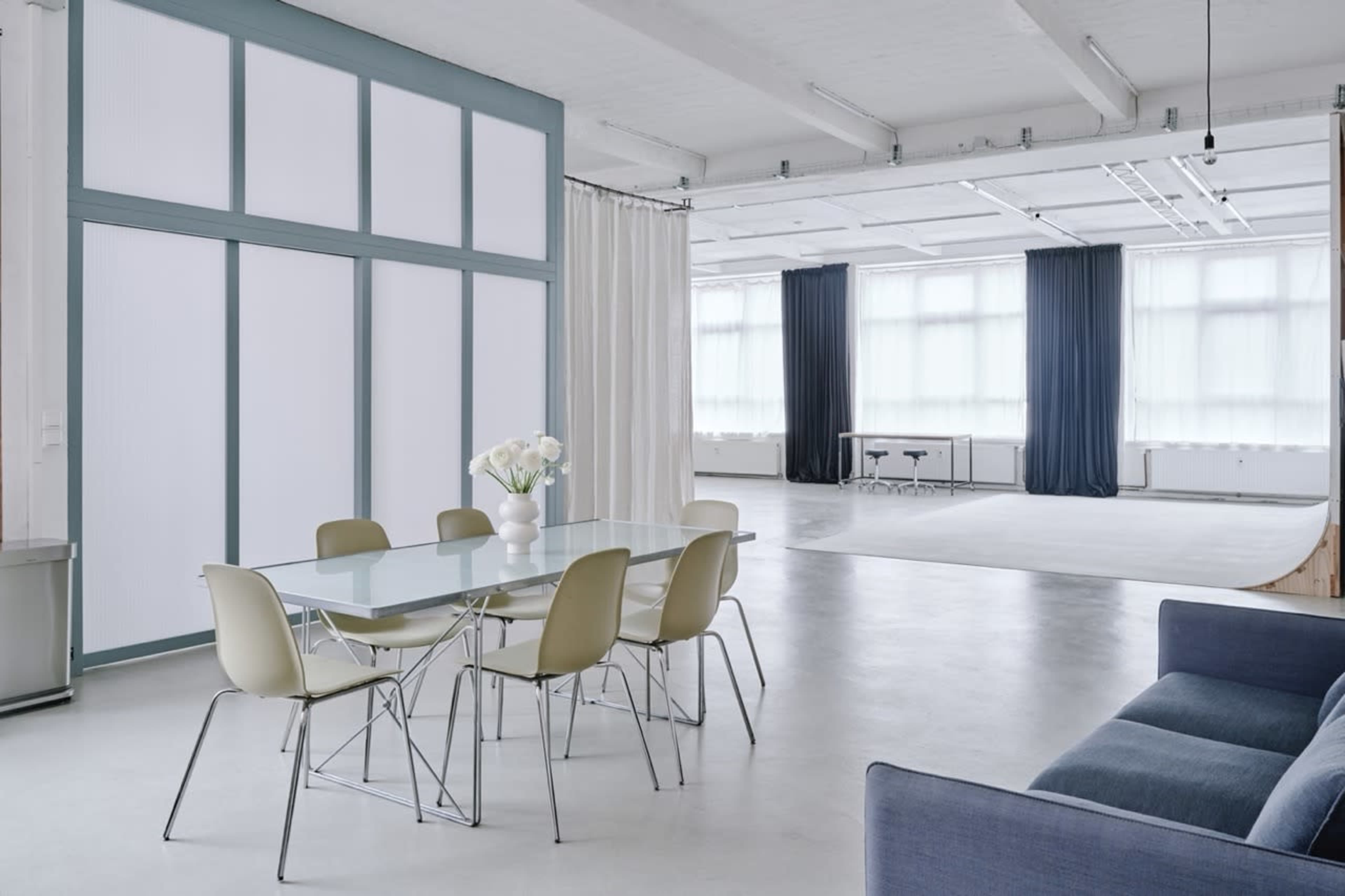 The image shows a modern, minimalistic interior with a dining area featuring a glass table and chairs, adjacent to a spacious, well-lit open area with large windows and light-colored walls.