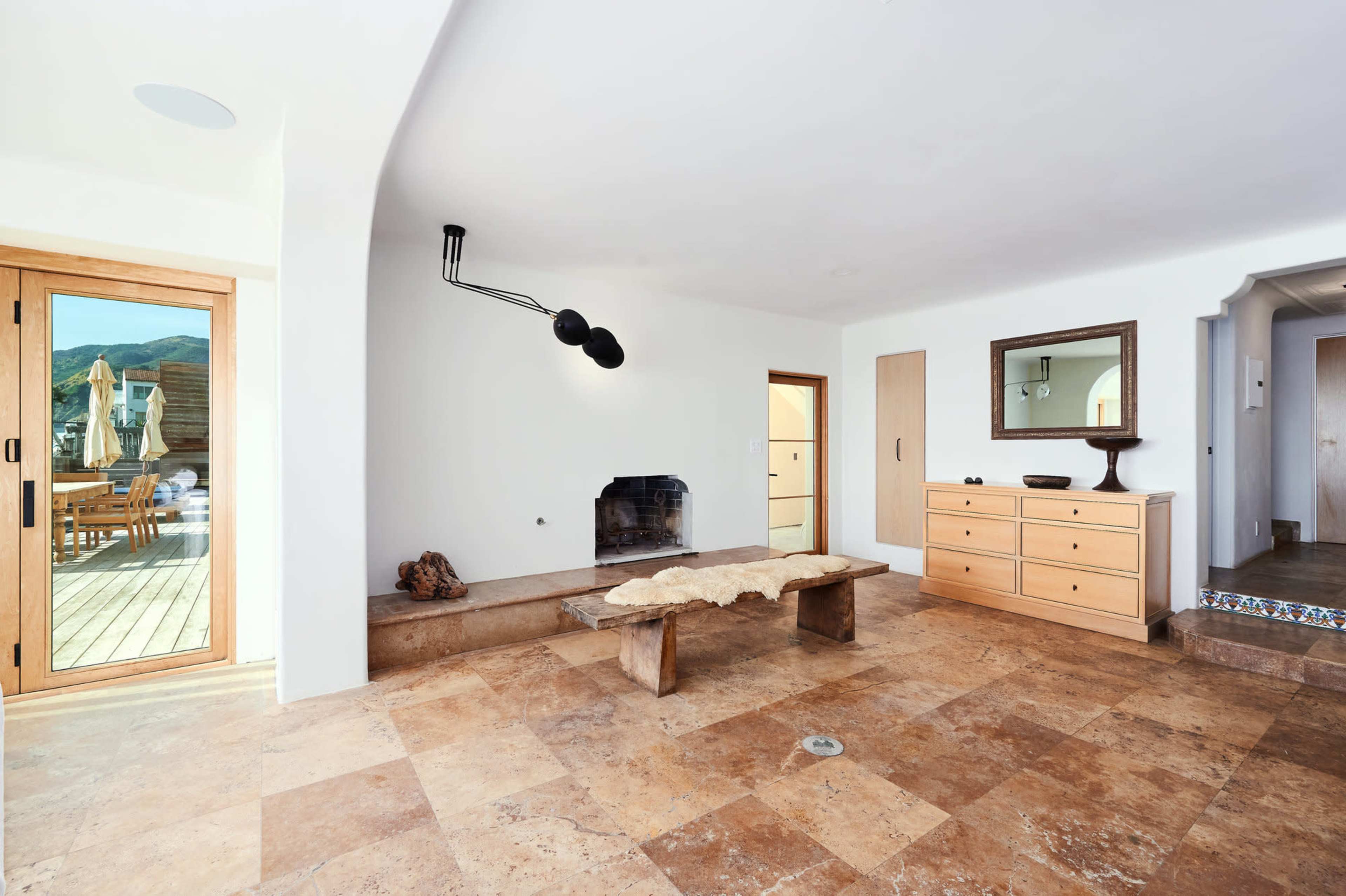 A minimalist living room with a wooden table, a fireplace, and a sliding door leading to an outdoor deck.