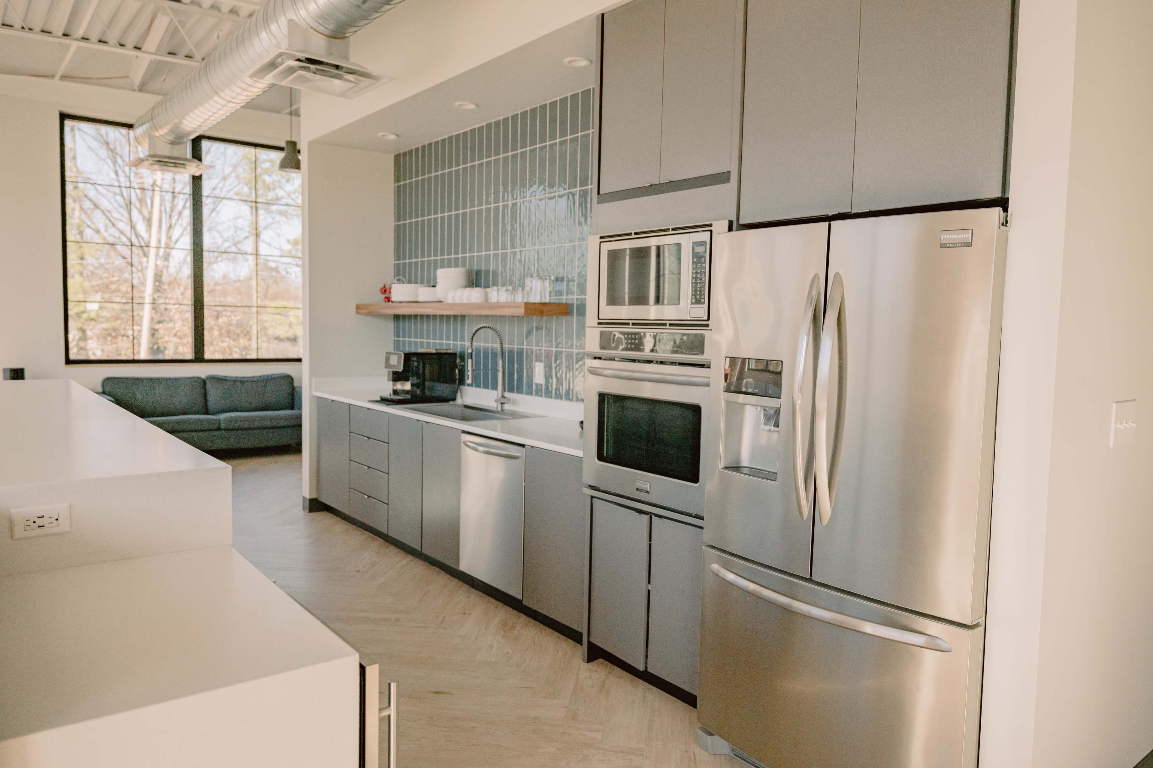 A modern kitchen featuring stainless steel appliances, grey cabinetry, and a window with a view of a couch and trees outside.