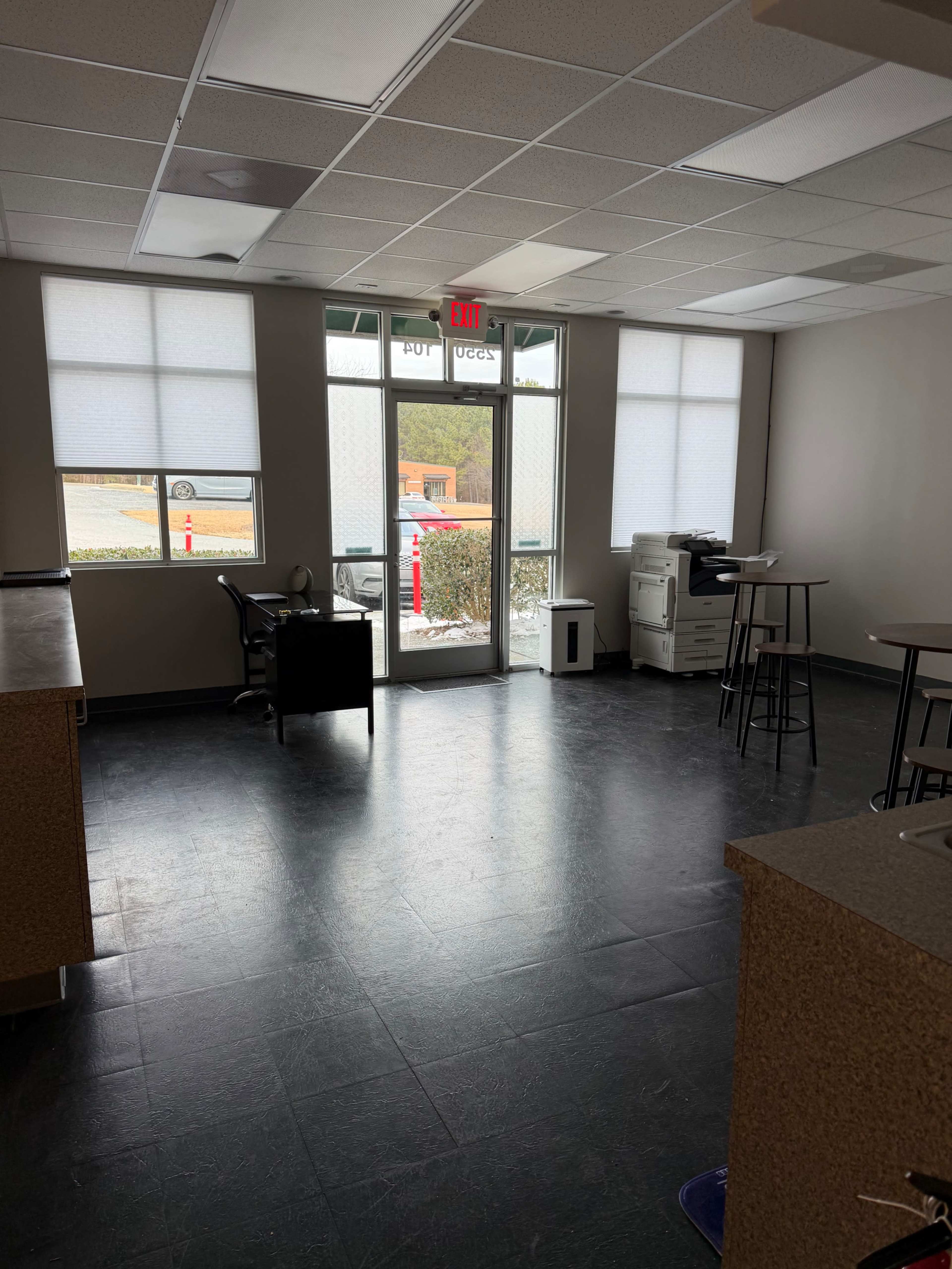 The image shows an empty office space with a front door, several windows, a few chairs, and a printer in the corner.