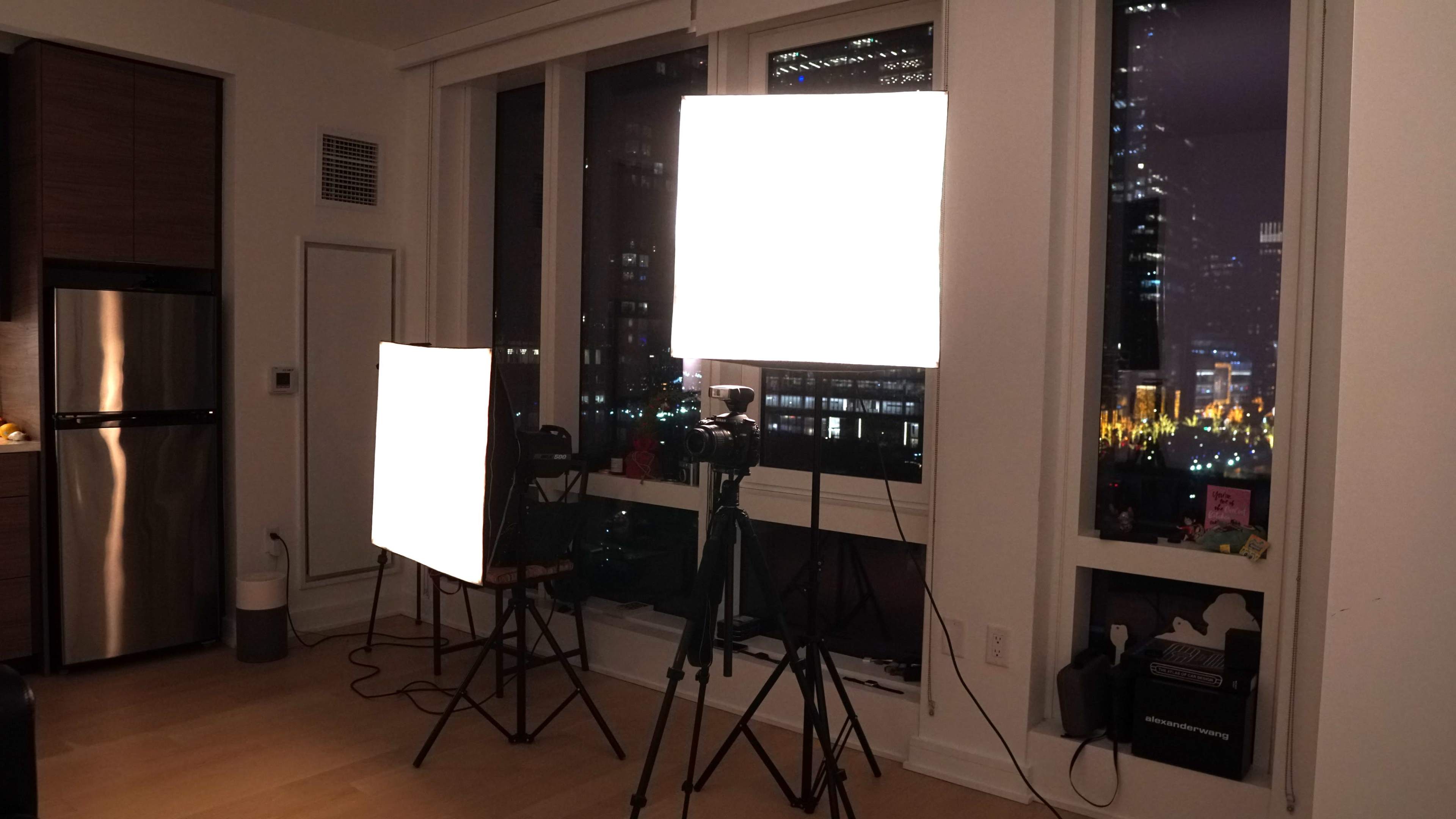 The image shows a modern room with two large softbox lights set up for photography, overlooking a cityscape at night through a window.