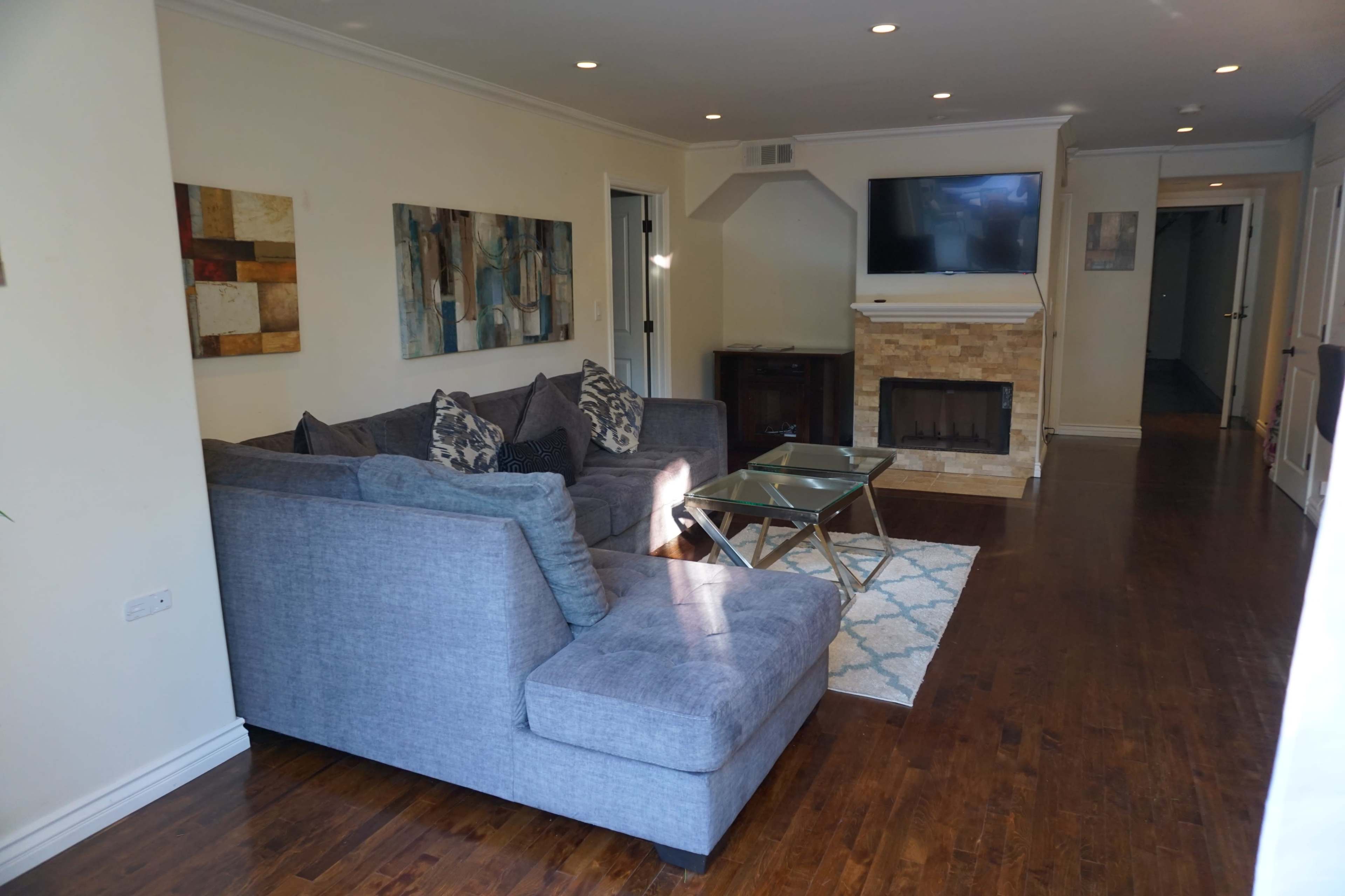 The image shows a living room with a gray sectional sofa, a glass coffee table, a stone fireplace, and a wall-mounted TV.