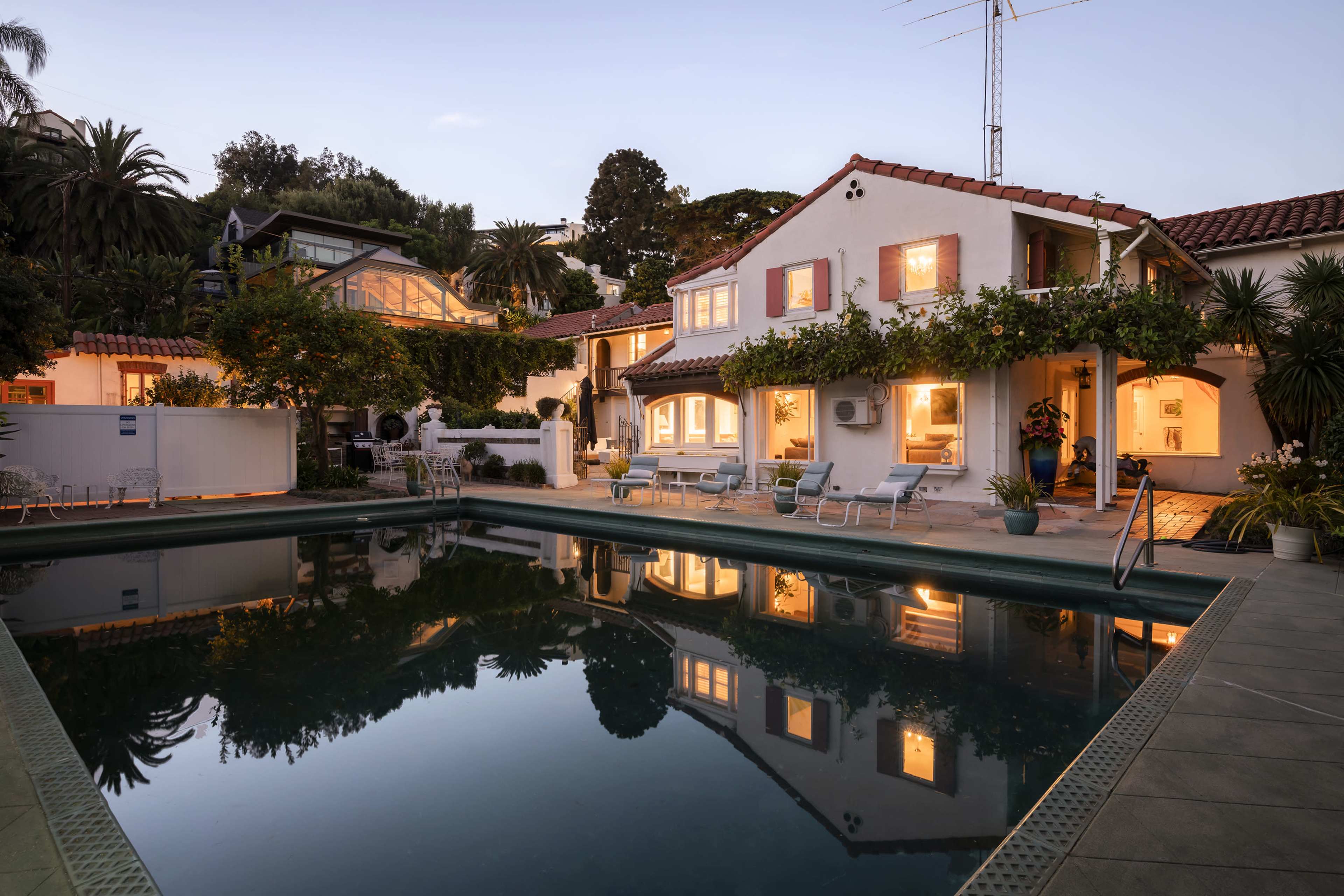 The image shows a tranquil swimming pool surrounded by lush greenery and illuminated buildings at dusk.