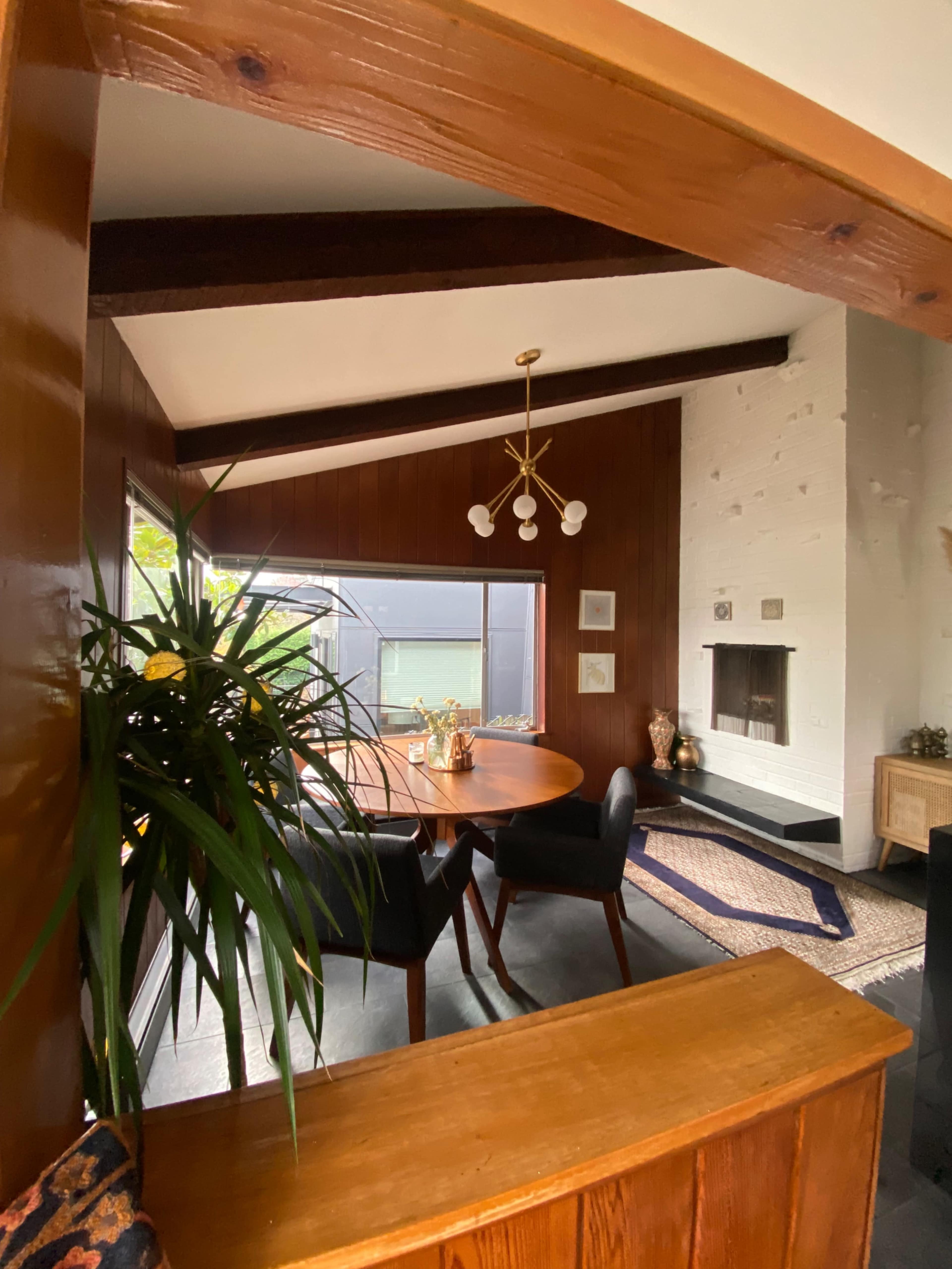 A dining area with a round table and black chairs, framed by wooden beams and a fireplace, with large windows providing natural light.