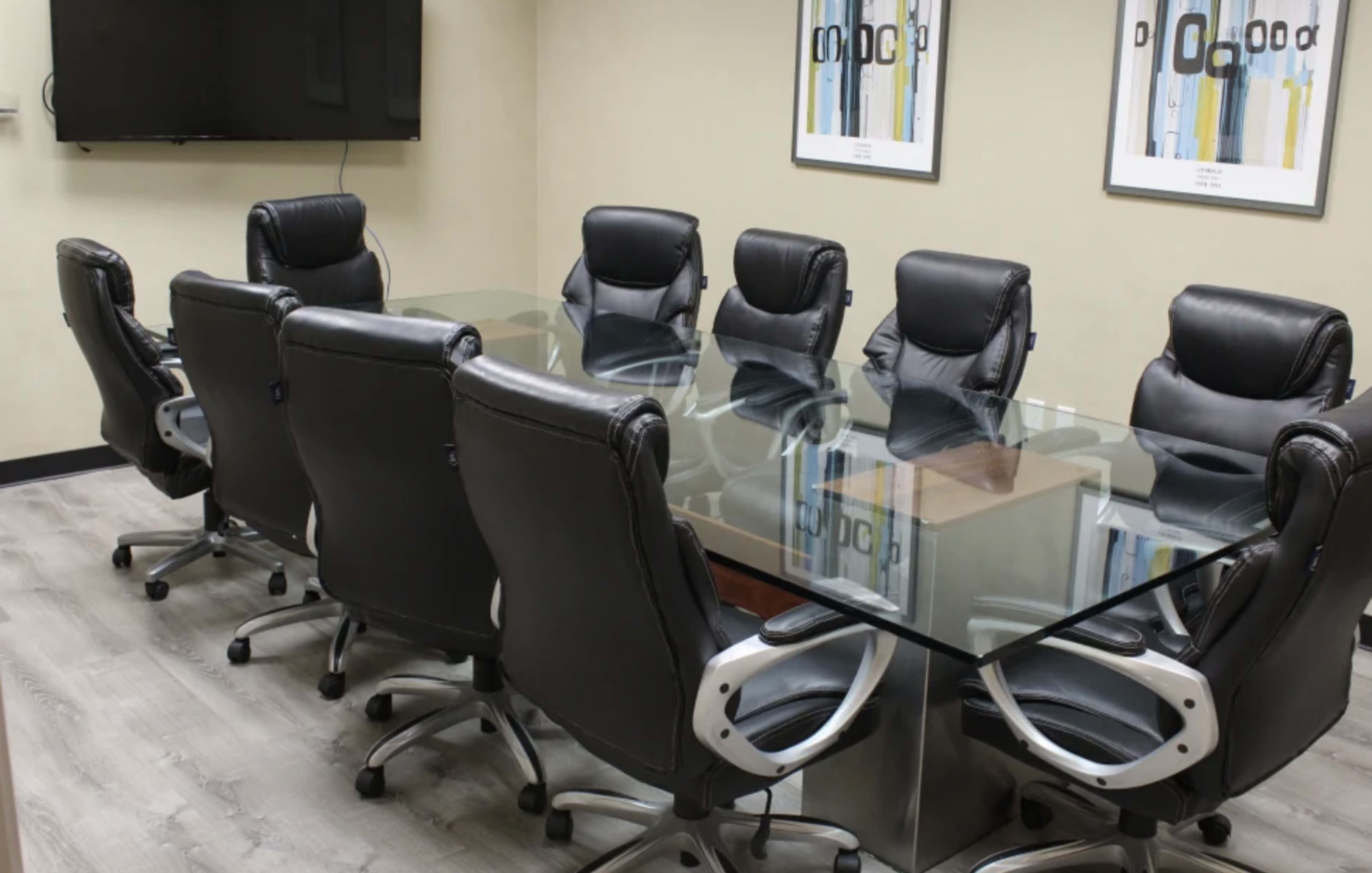 A conference room is set up with a glass table surrounded by eight black leather chairs, with a television mounted on the wall and framed artwork nearby.