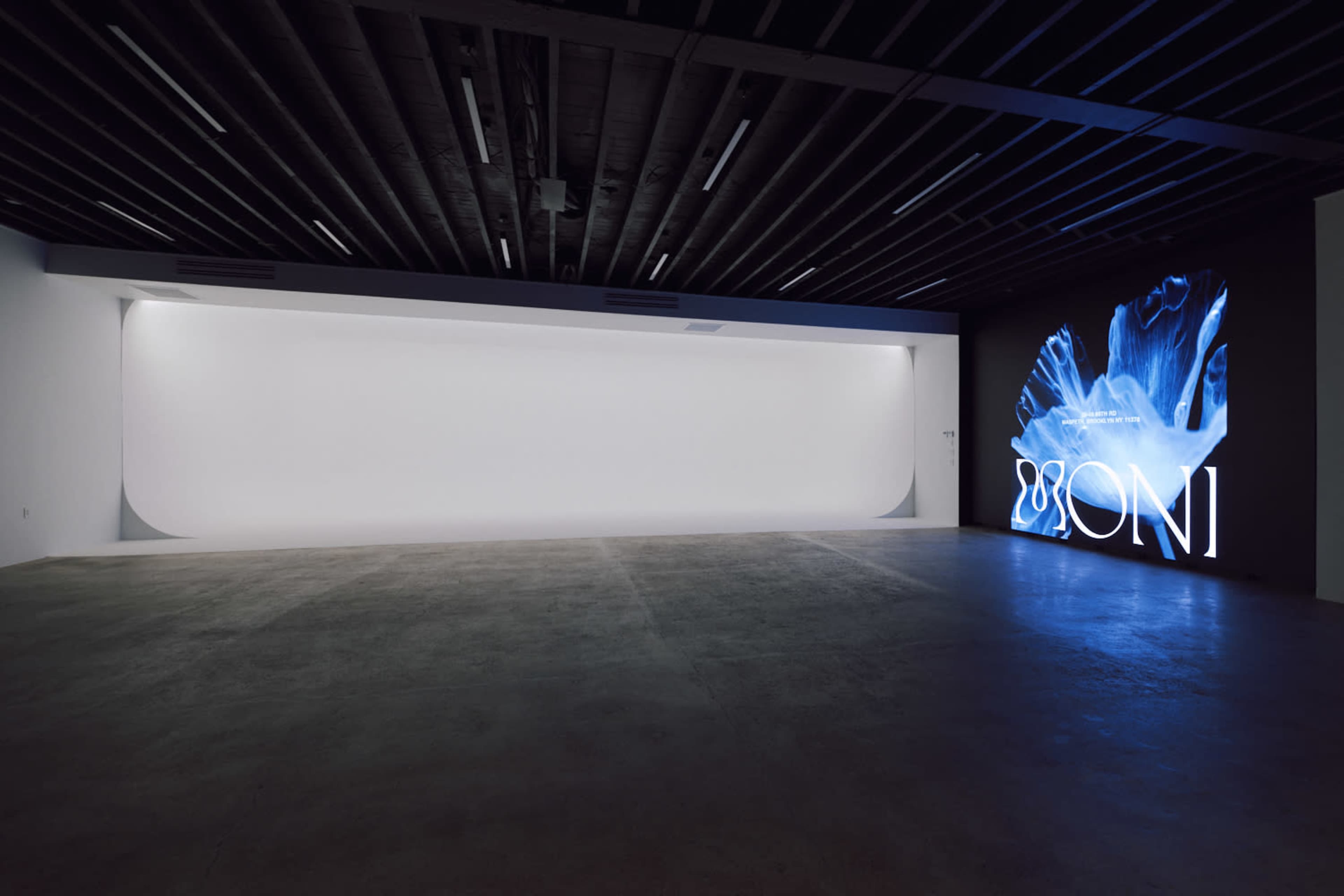 The image shows an empty art gallery space with a large, illuminated digital display on one wall and a blank white area opposite it.