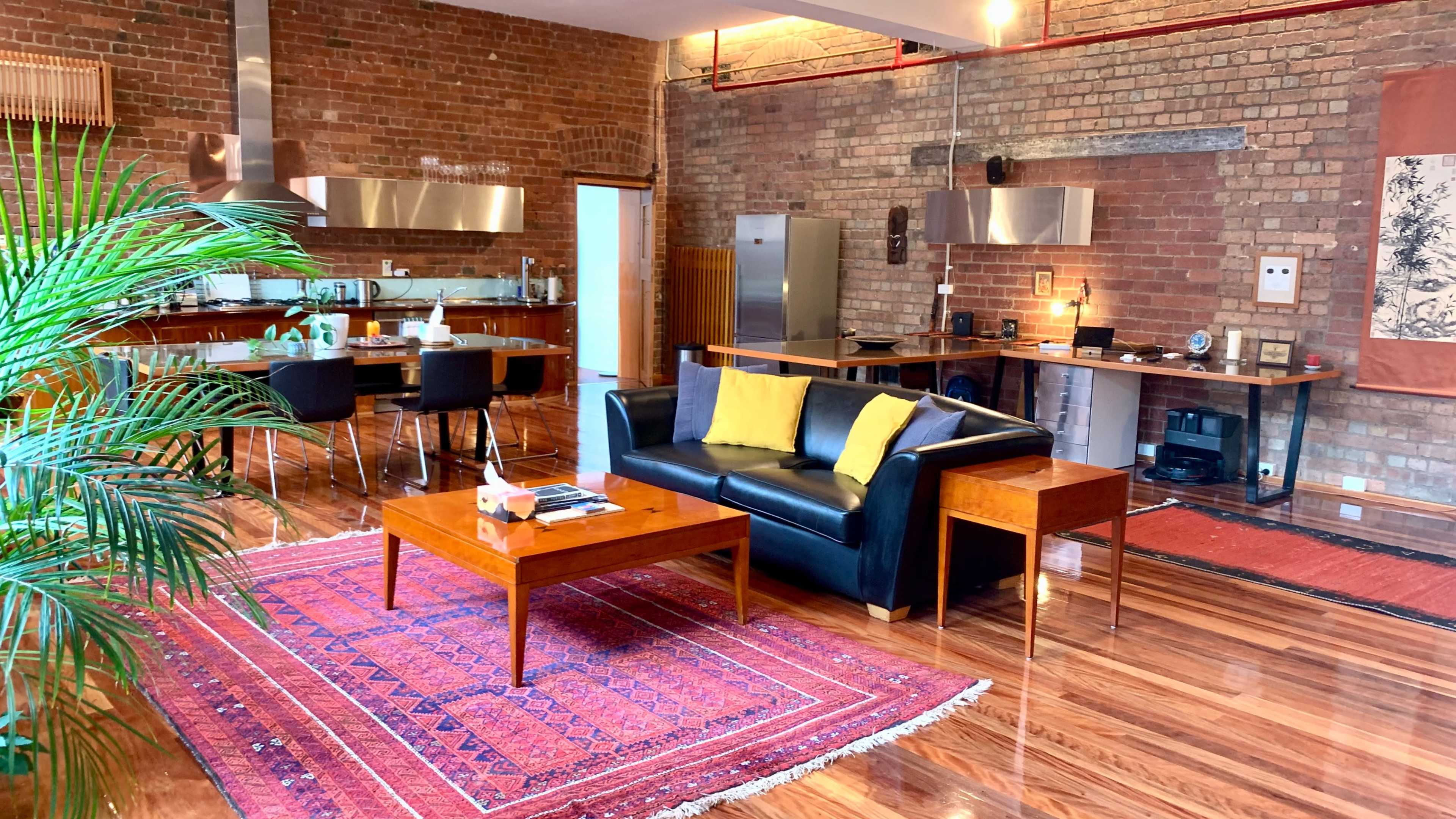 A modern, open-concept living space featuring a black leather couch, a wooden coffee table, and a kitchen area with stainless steel appliances, all set against exposed brick walls and hardwood flooring.