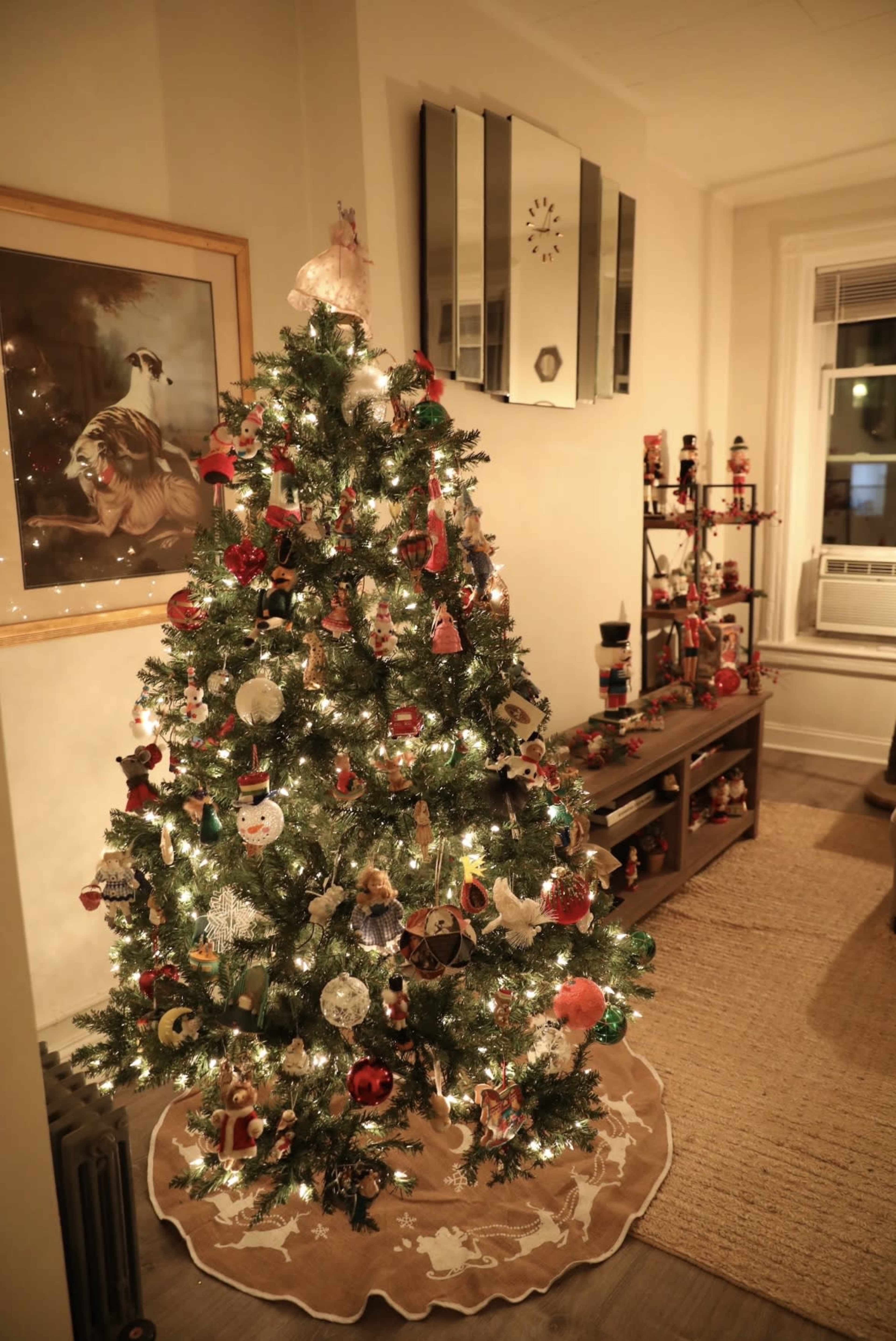 A decorated Christmas tree with lights and ornaments stands in a cozy room beside a shelf of festive figures.