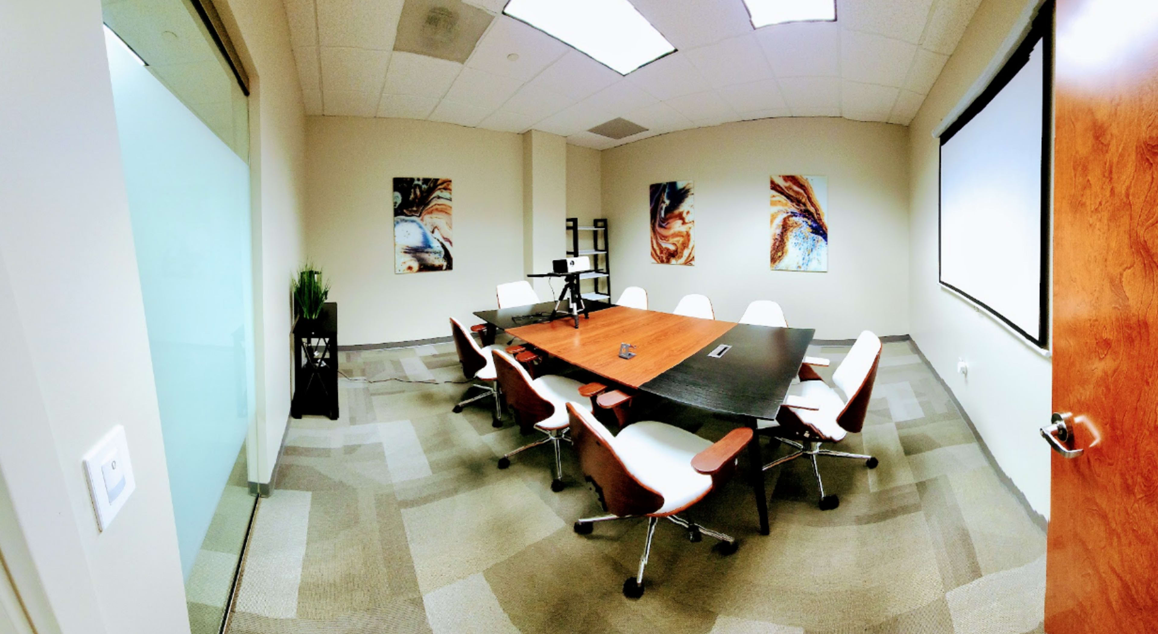 A modern conference room with a large wooden table surrounded by white chairs, featuring artwork on the walls and a projector screen.