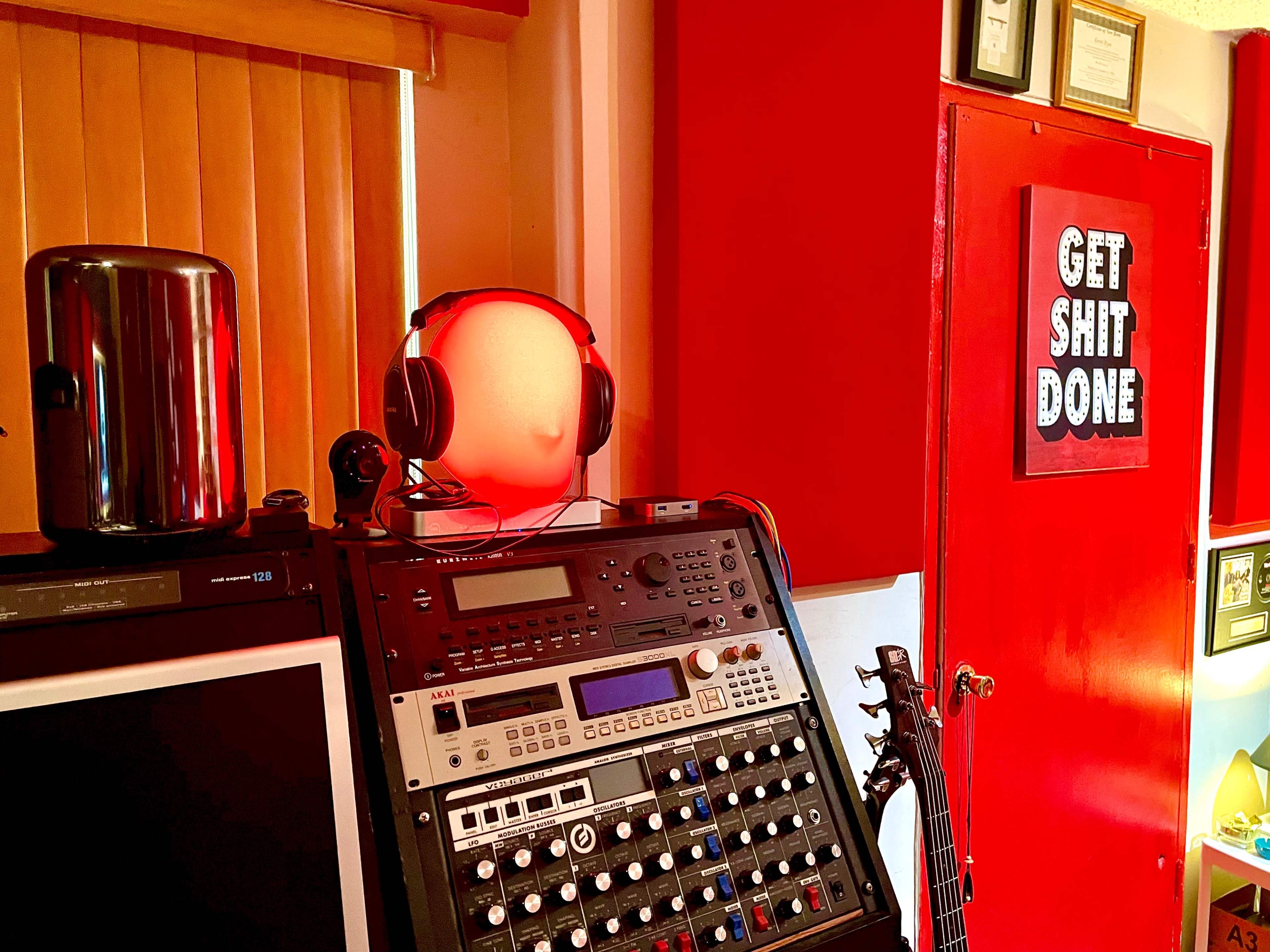A music production setup featuring a mixing console, a computer monitor, headphones on a light fixture, and a red door with a motivational sign.