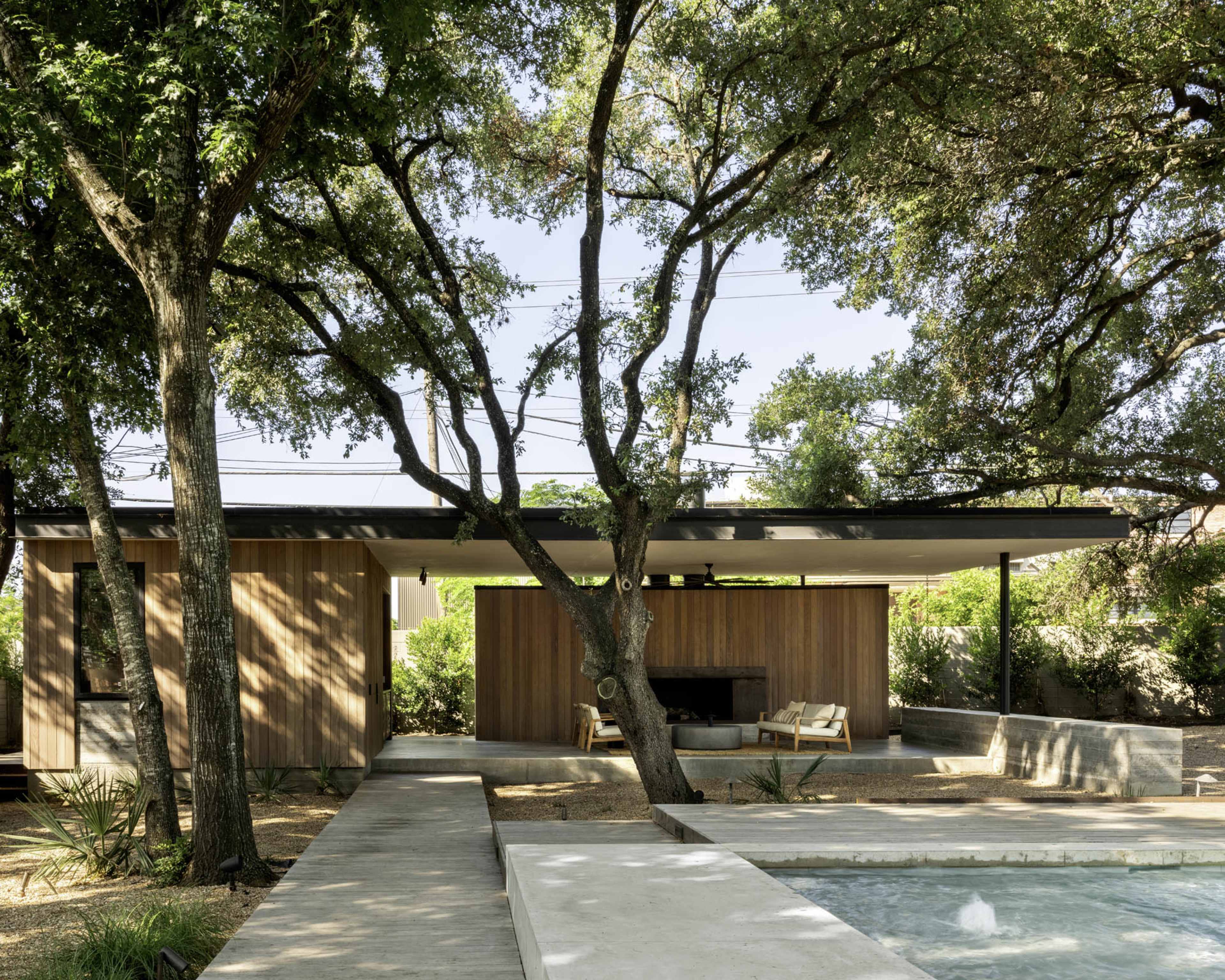 A modern house with wooden siding and a covered patio is surrounded by trees and features a concrete pathway leading to a small pool.