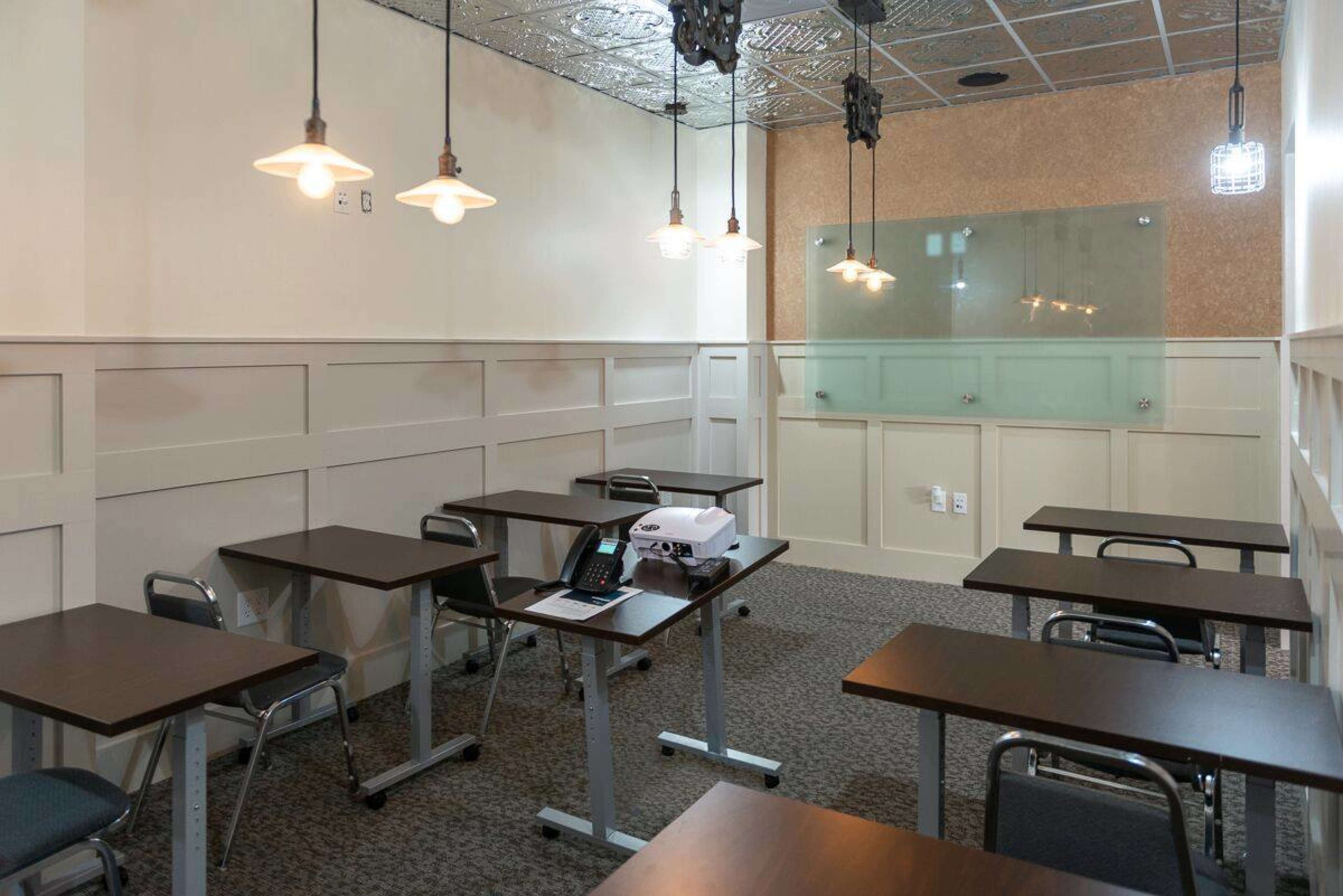 A small, well-lit classroom features several wooden tables and chairs, with a projector and telephone on one table.