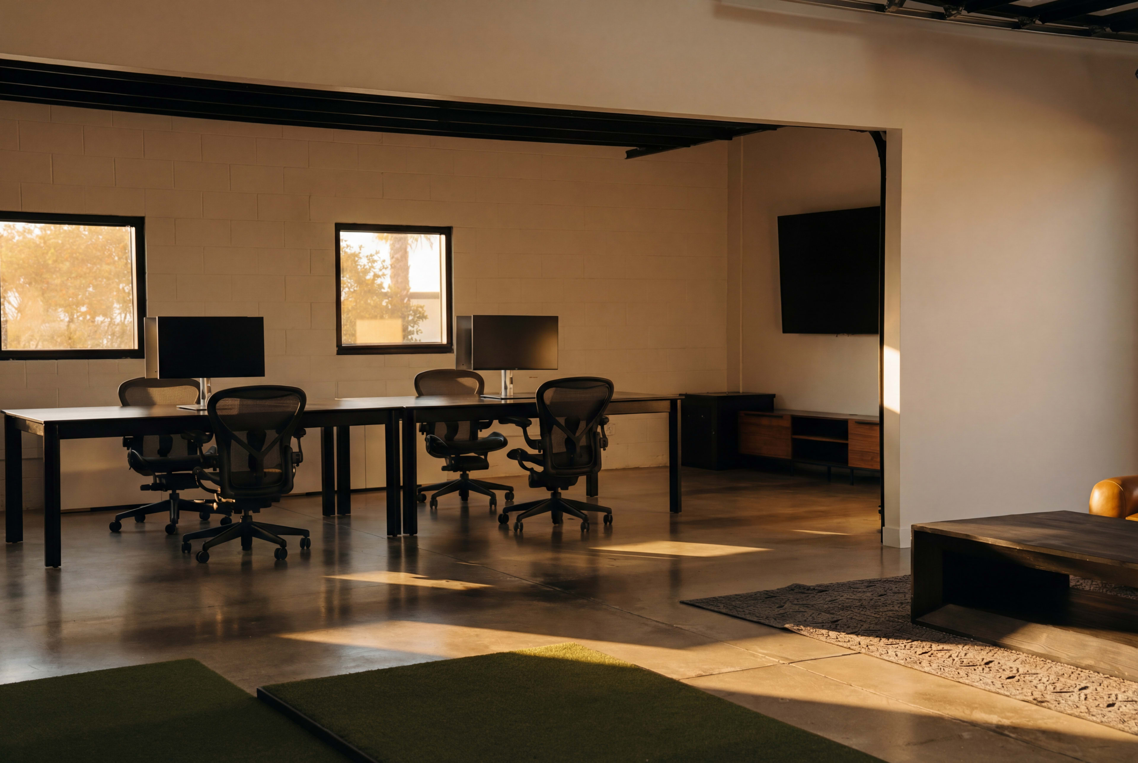 The image shows a modern office space featuring several workstations with ergonomic chairs, large windows allowing natural light in, and sleek furniture.