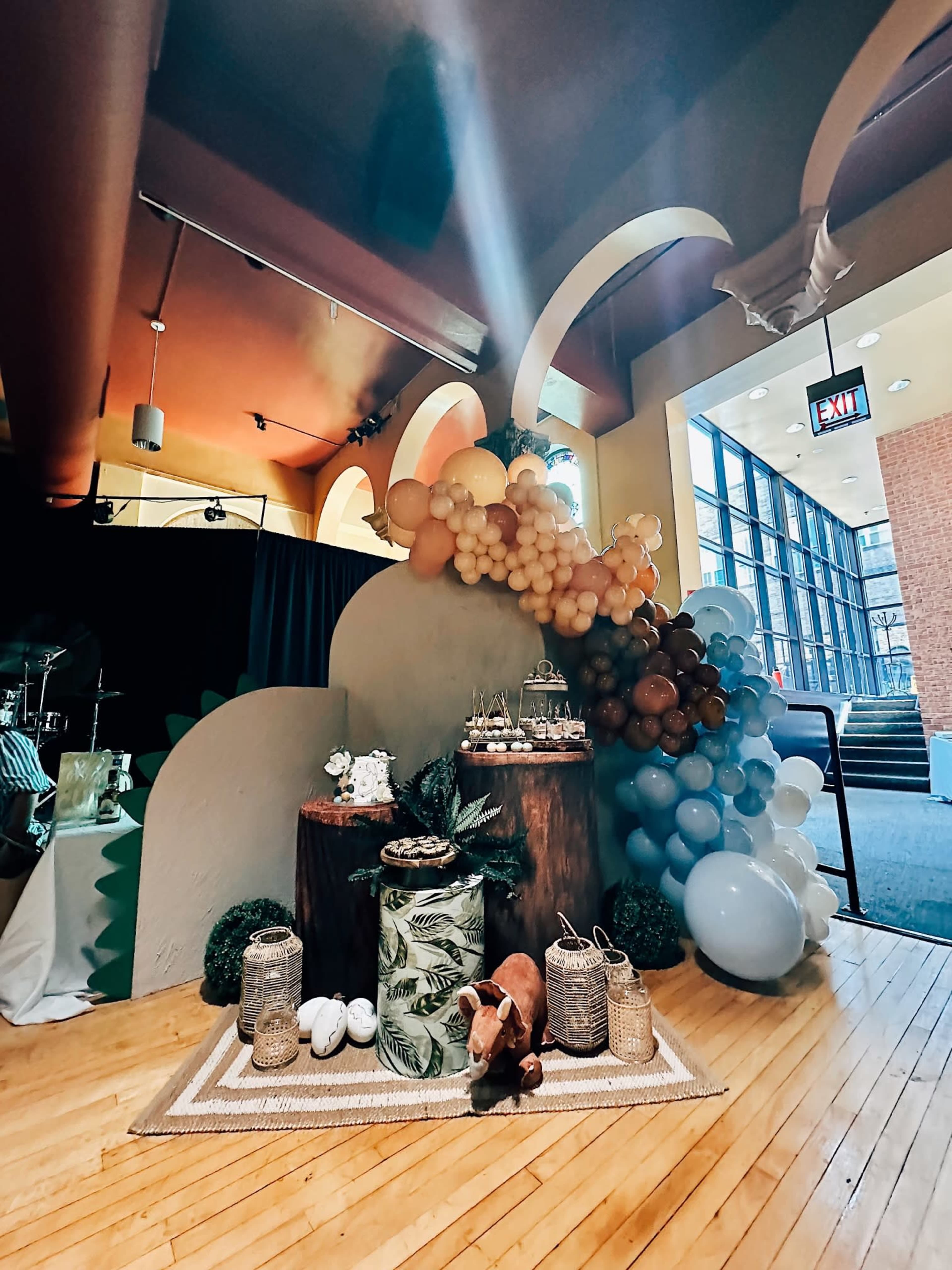A decorated event space featuring a backdrop of balloons, wooden displays, and a table with pastries and ornamental items.
