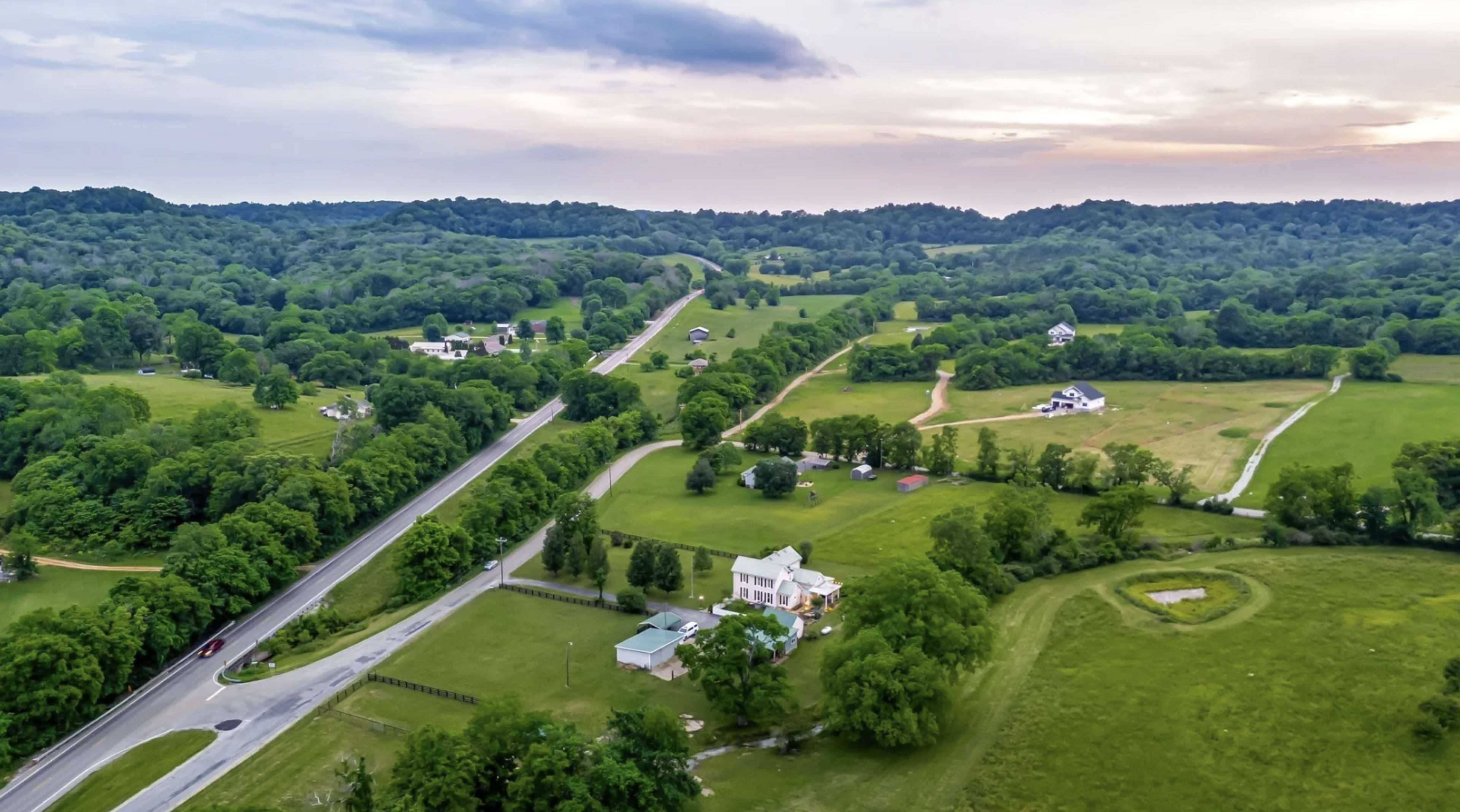 The image shows a rural landscape with a winding road, scattered houses, and green fields under a partly cloudy sky.