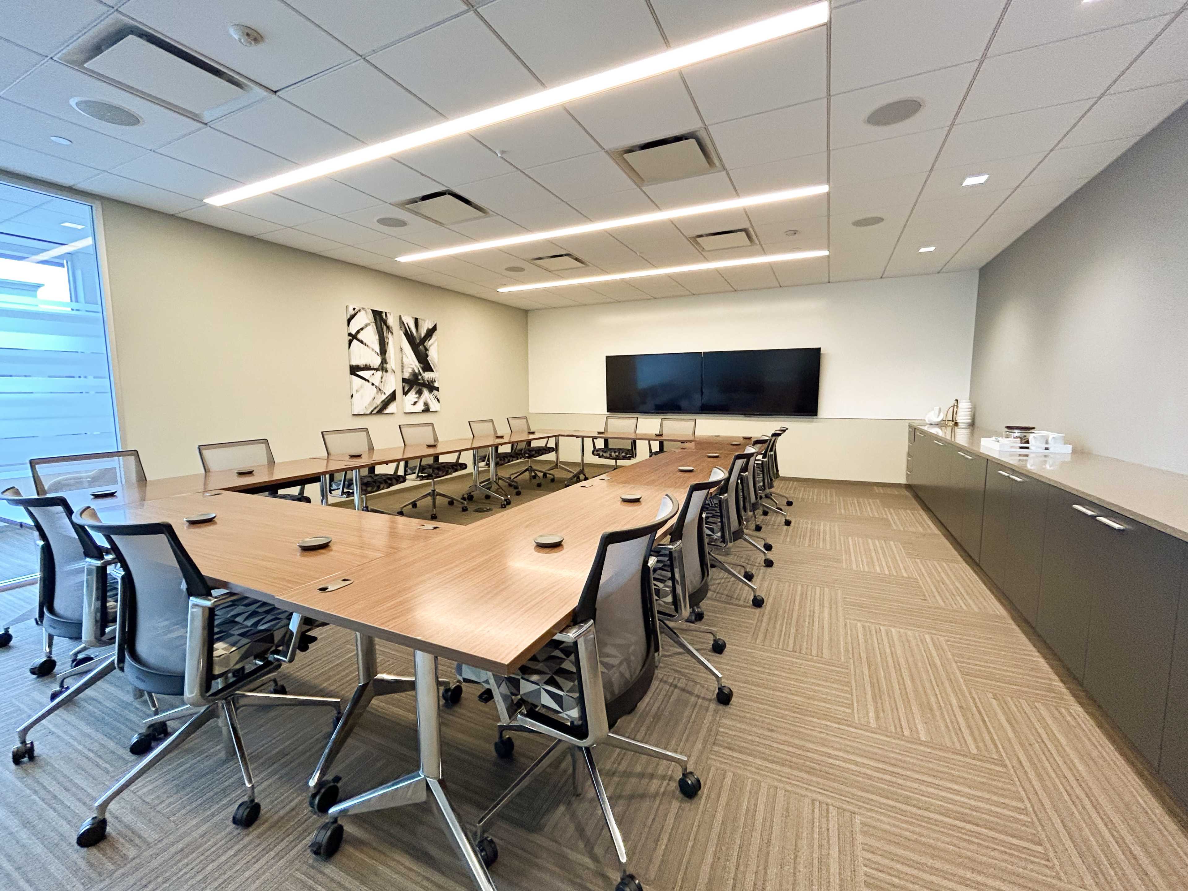 A modern conference room features a large oval table surrounded by ergonomic chairs, with a wall-mounted screen and a side cabinet for refreshments.