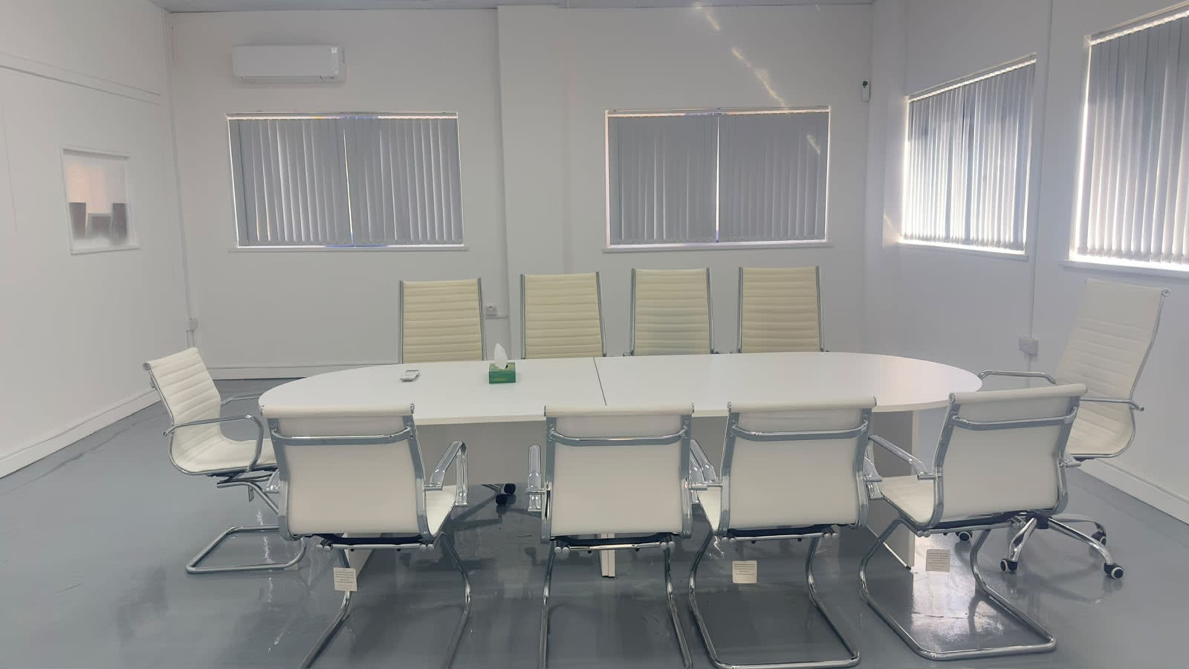 A modern conference room features a long white table surrounded by eight white ergonomic chairs, with large windows allowing natural light to enter.