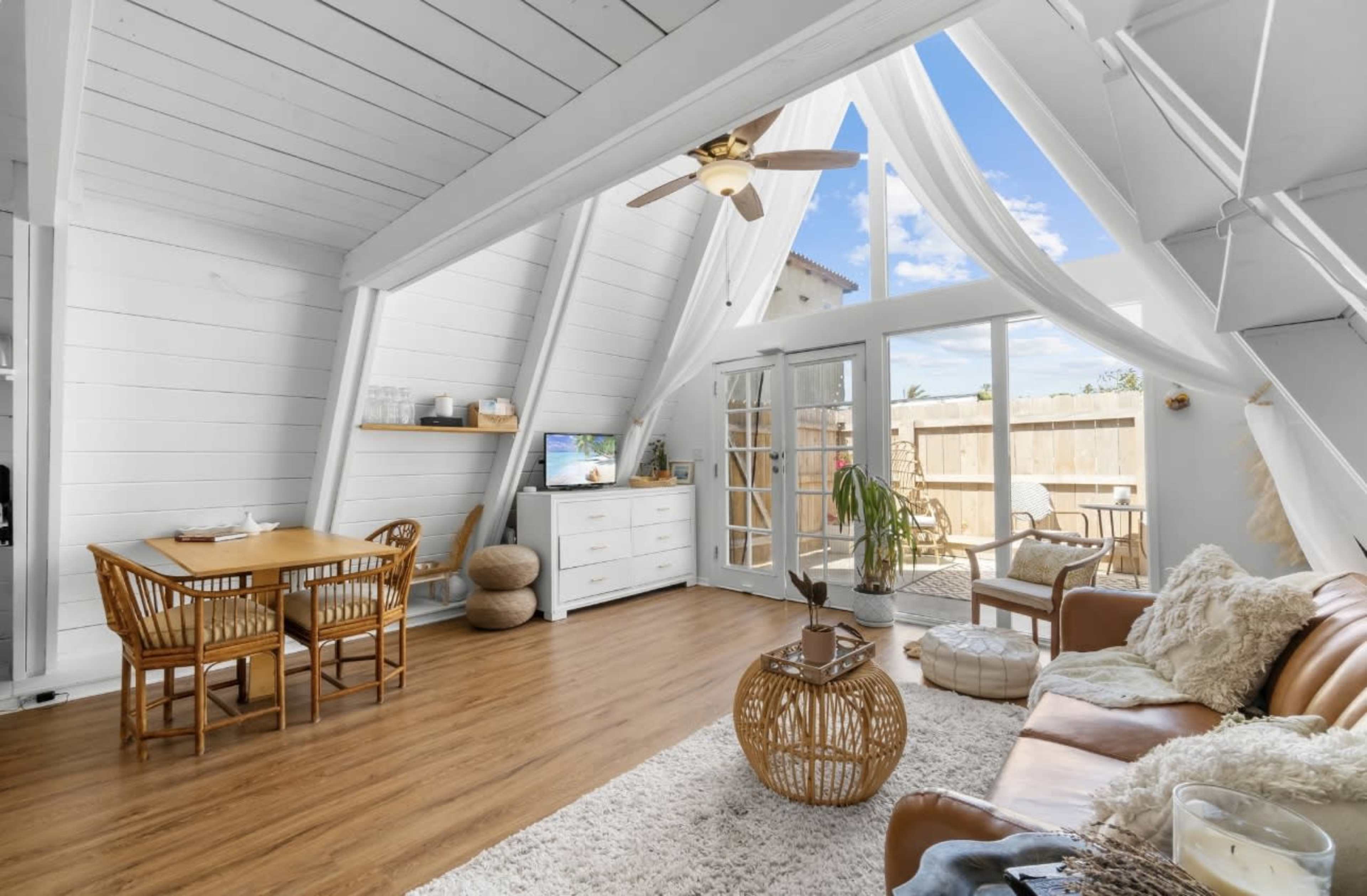 A bright, airy living space features a wooden dining table, a cozy seating area, and large windows allowing natural light to flood the room.