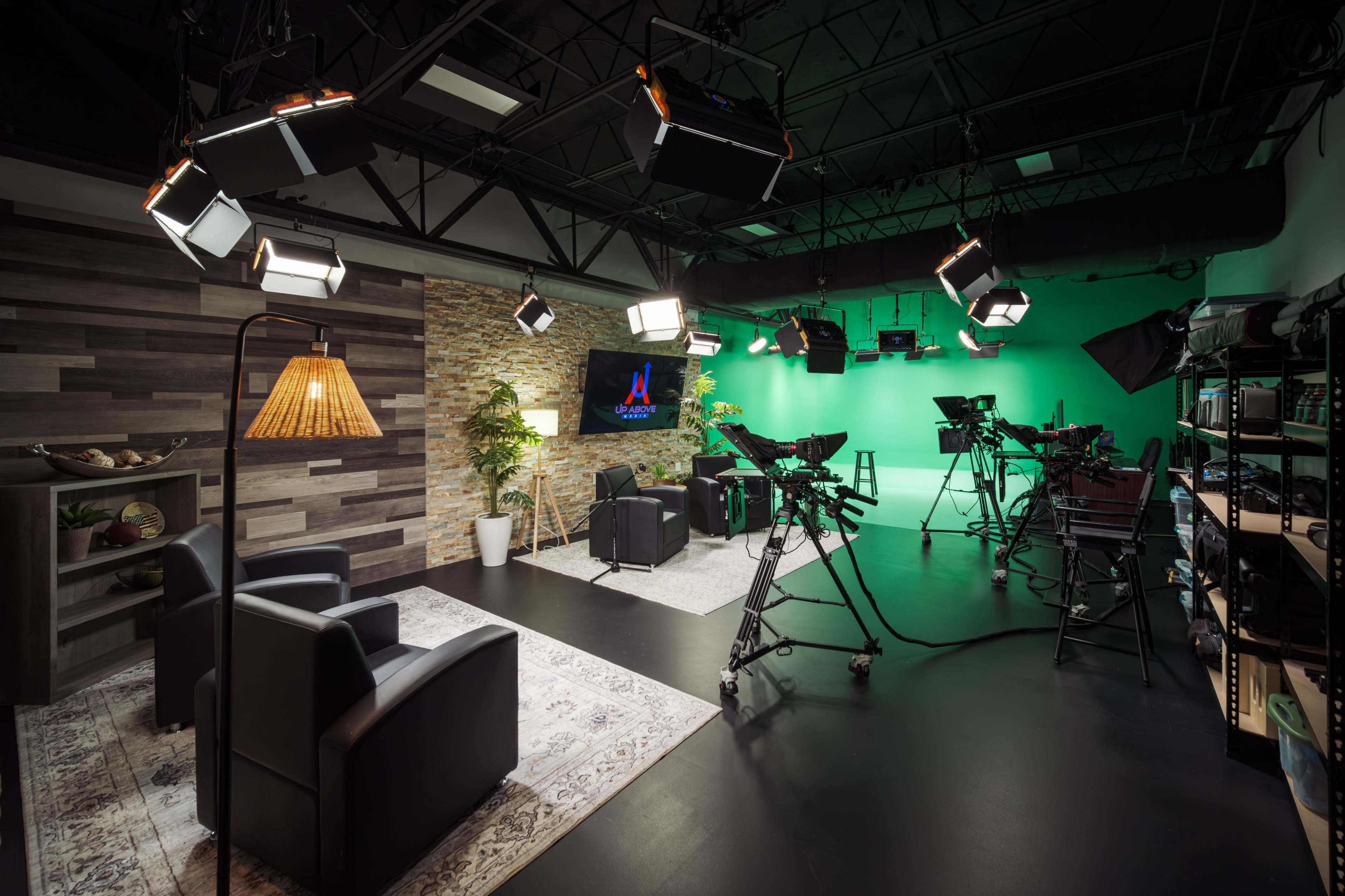 The image shows a modern studio setup with two black chairs, lighting equipment, and cameras arranged around a green screen and a decorative stone wall.