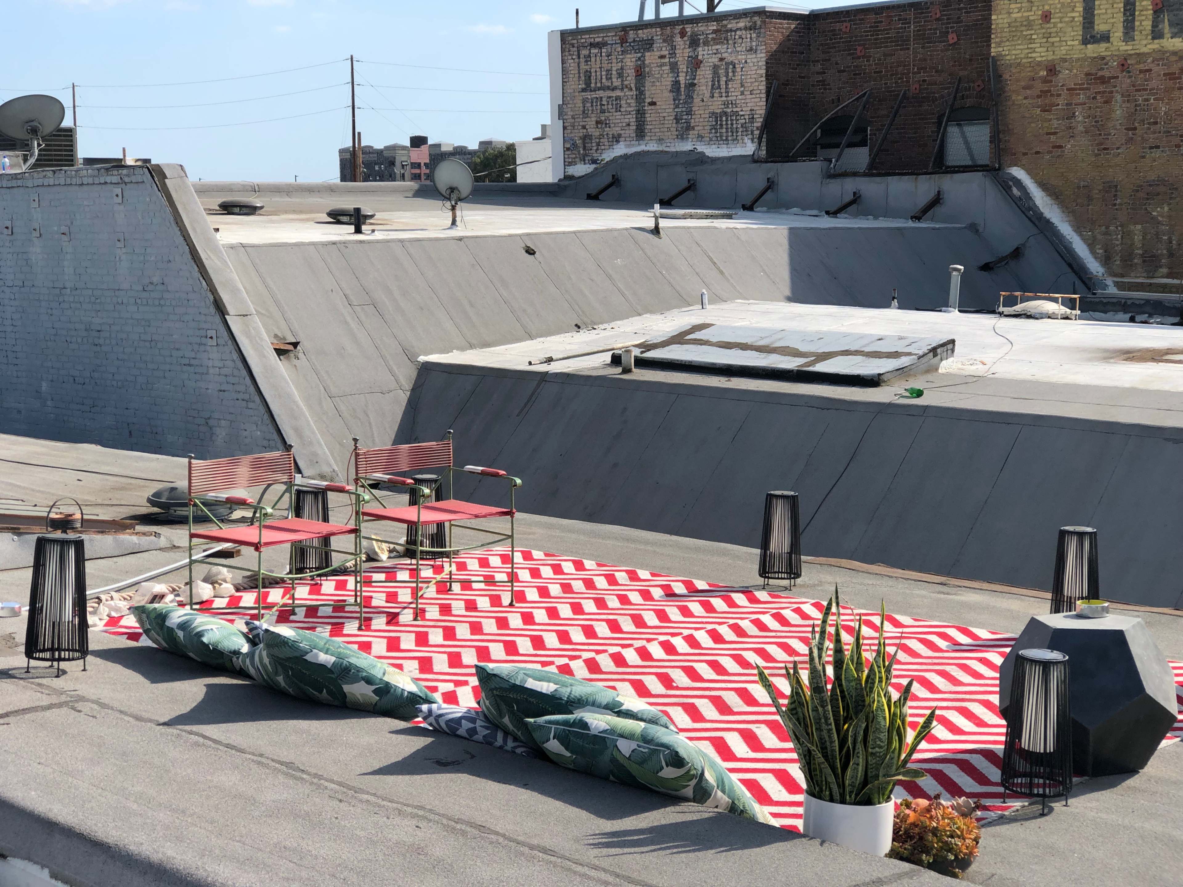 A rooftop area featuring two metal chairs, a striped rug, several large cushions, lanterns, and a potted plant.