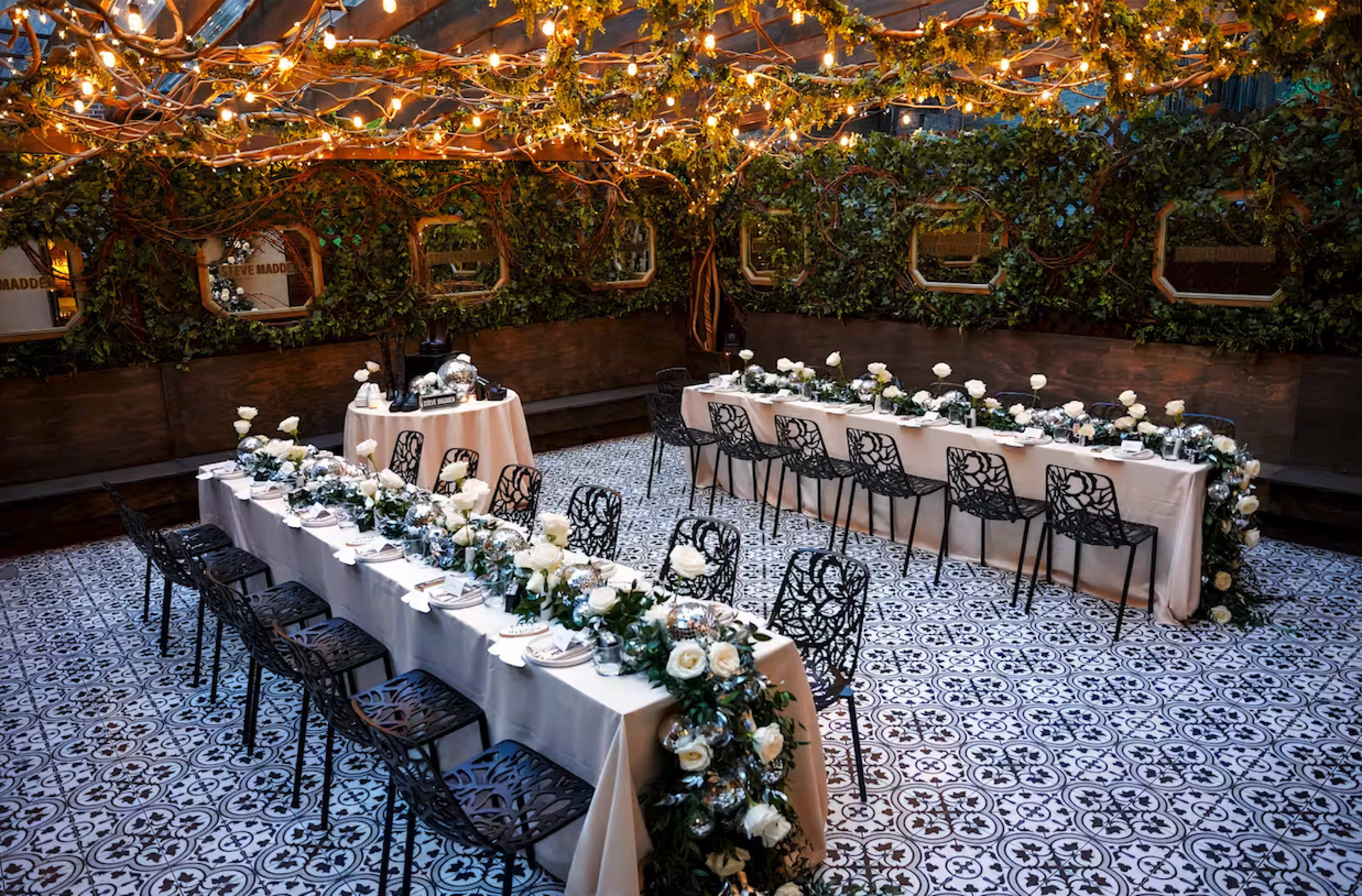 A dining area is arranged with long tables adorned with white tablecloths and floral centerpieces under warm string lights and green foliage.