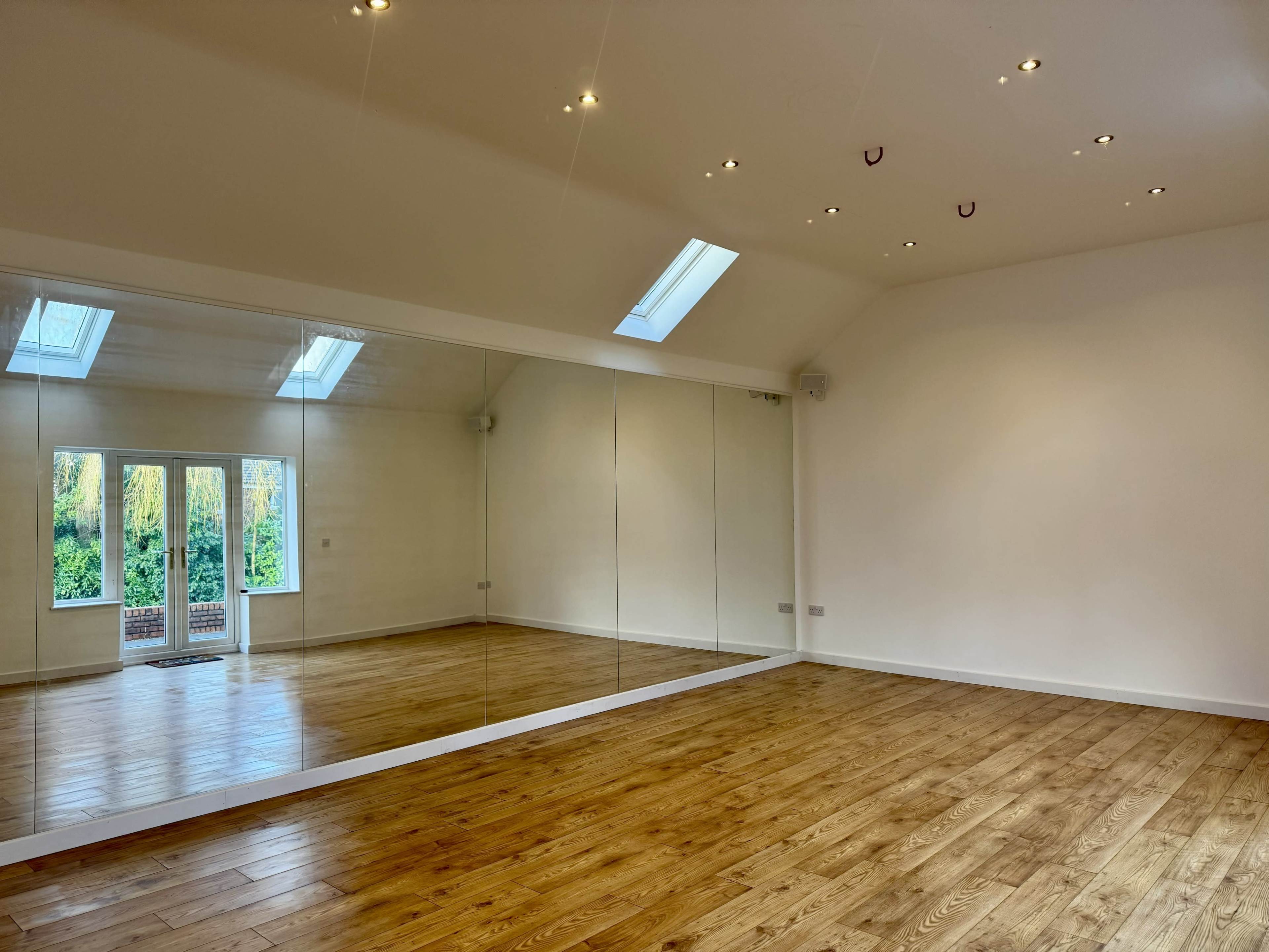 The image shows a spacious room with wooden flooring, a large mirror along one wall, and skylights providing natural light.
