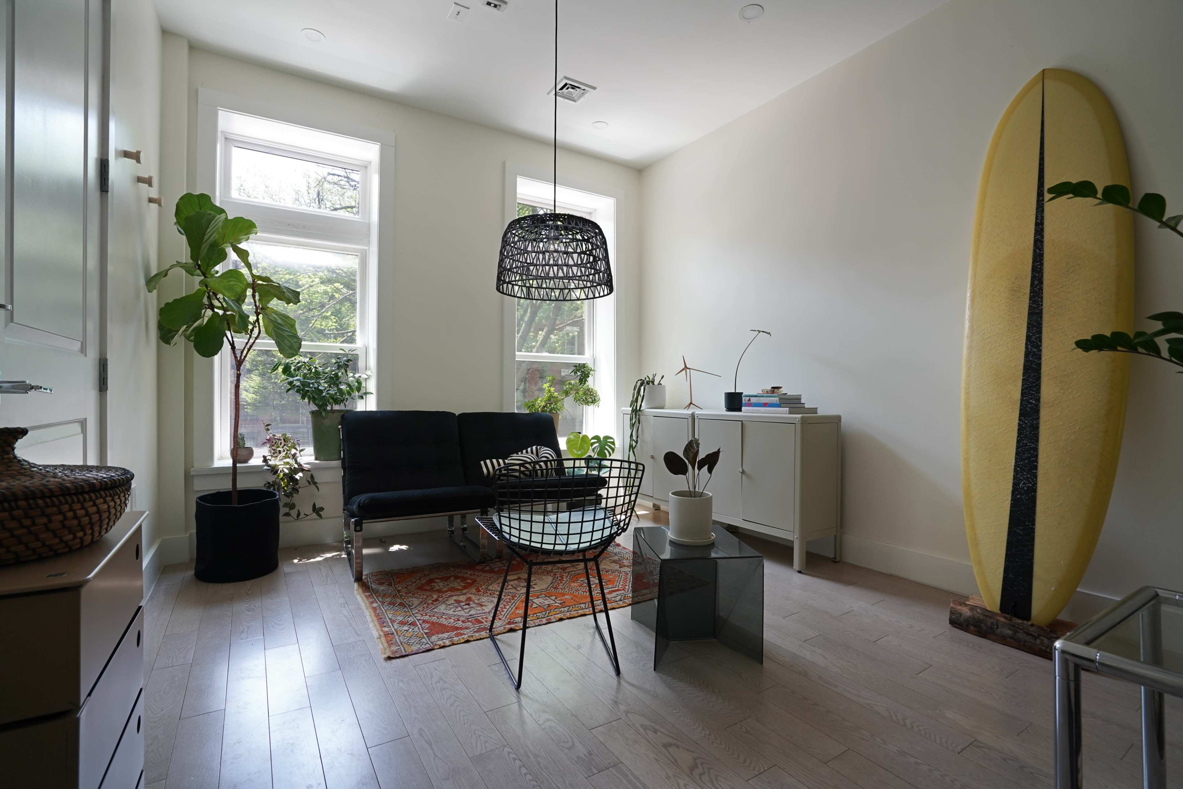 The room features a minimalist design with a black wire chair, a coffee table, a yellow surfboard leaning against the wall, and various potted plants.