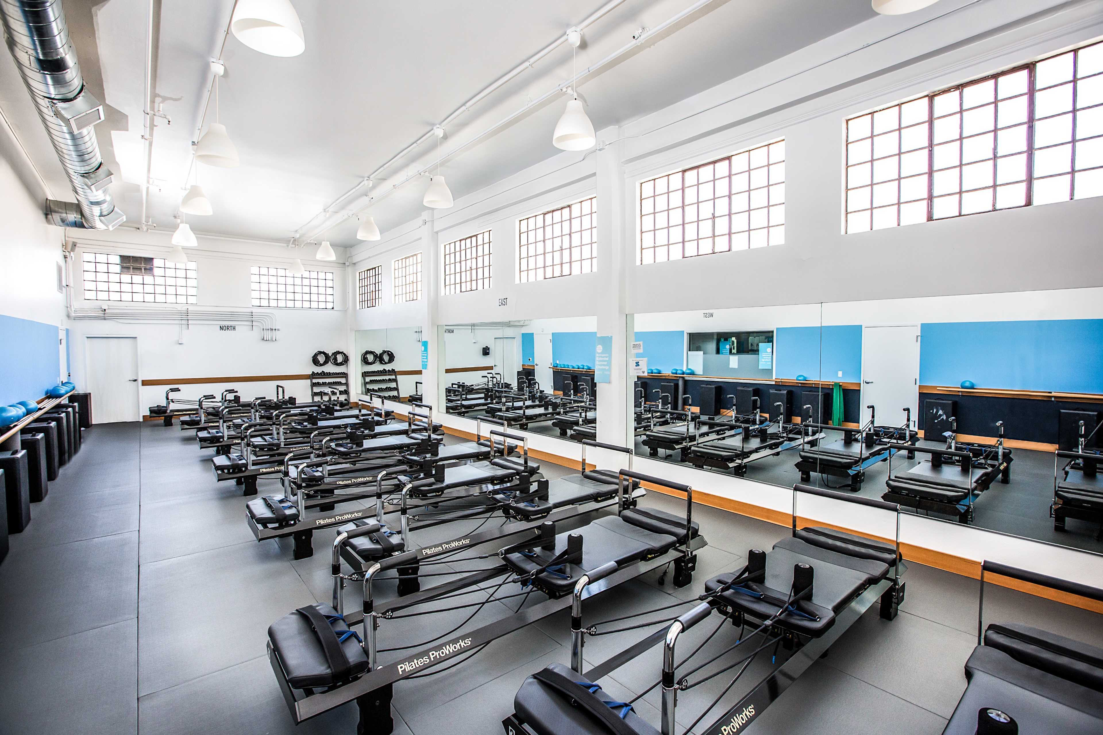 A bright, spacious Pilates studio filled with reformer machines arranged in neat rows, surrounded by large windows and mirrors.