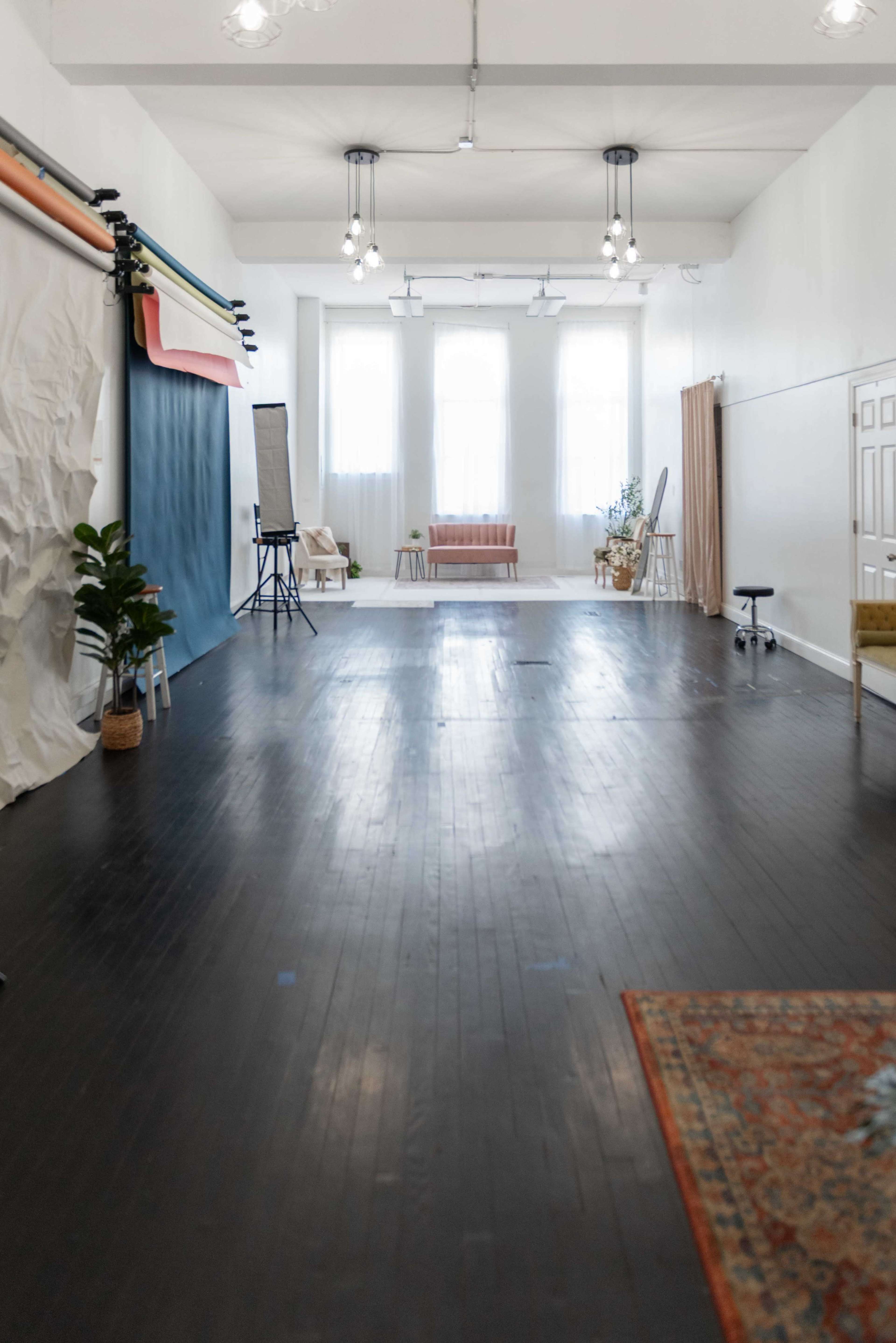 The image features a spacious, well-lit studio with hardwood floors, large windows, and various seating arrangements along the sides.