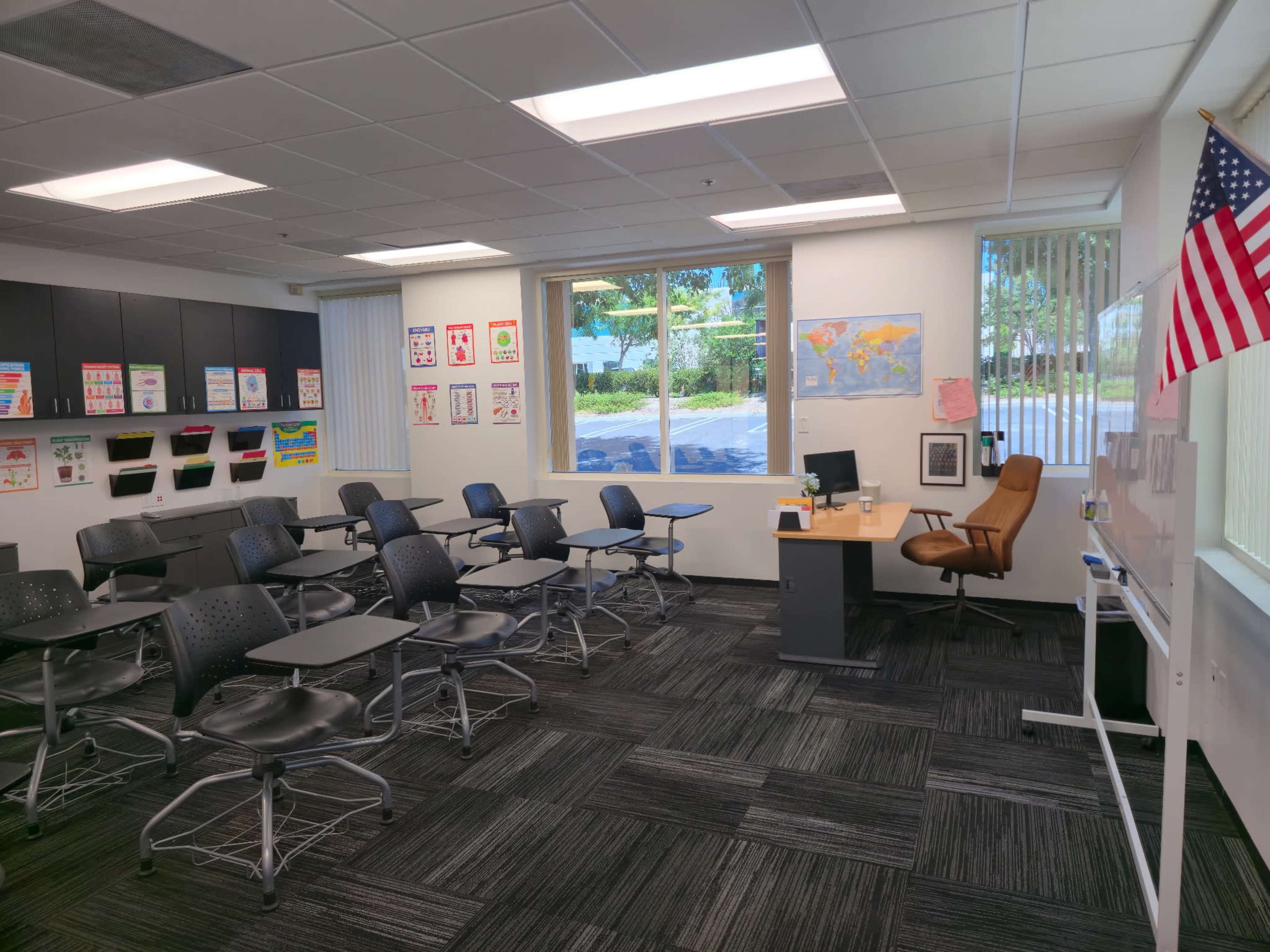 A classroom features rows of black chairs and desks, large windows letting in natural light, a map on the wall, and an American flag hanging by the window.