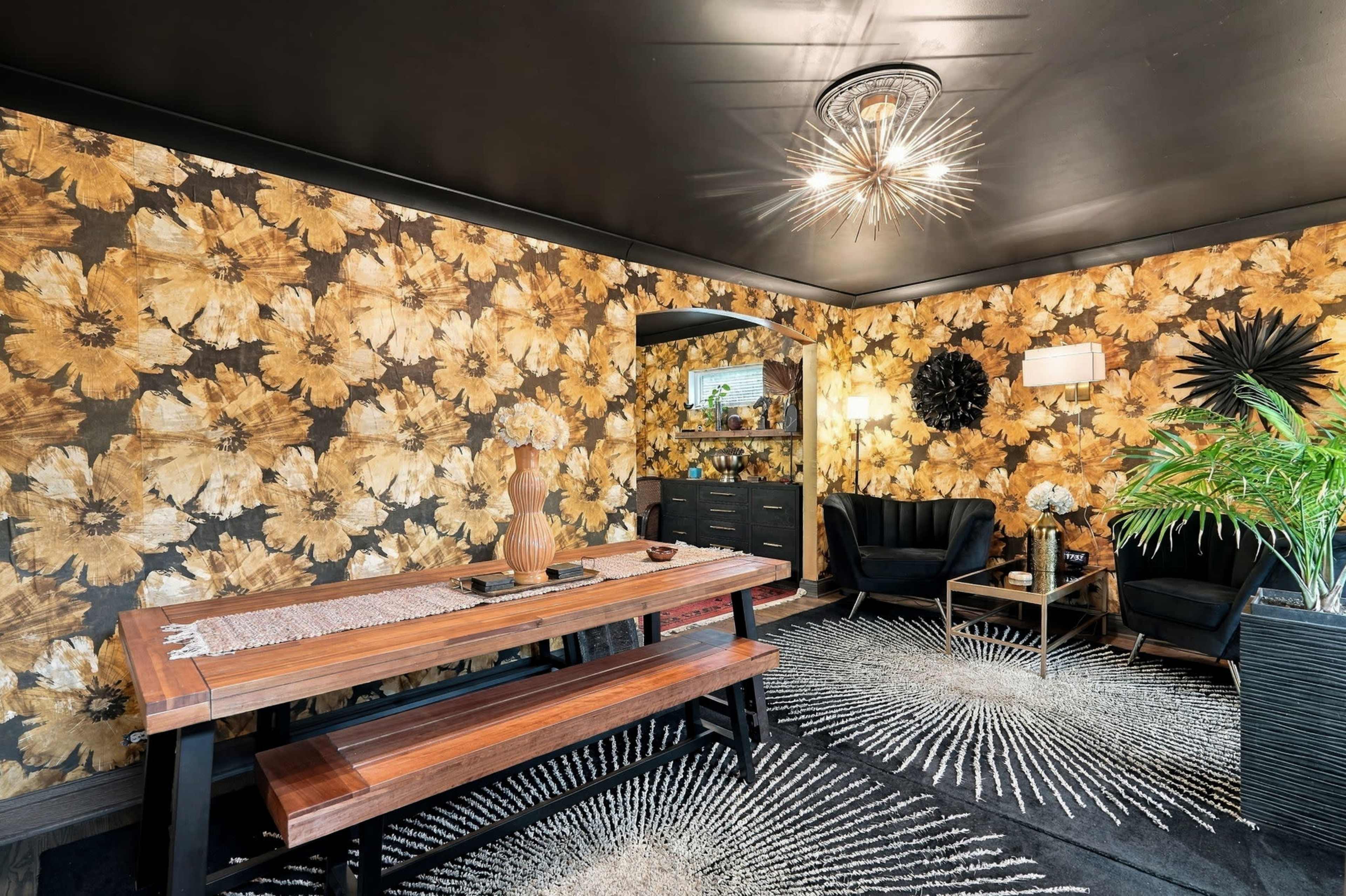 The scene features a dining area with a wooden table and chairs, surrounded by floral-patterned wallpaper, a large area rug, and modern decorative elements.
